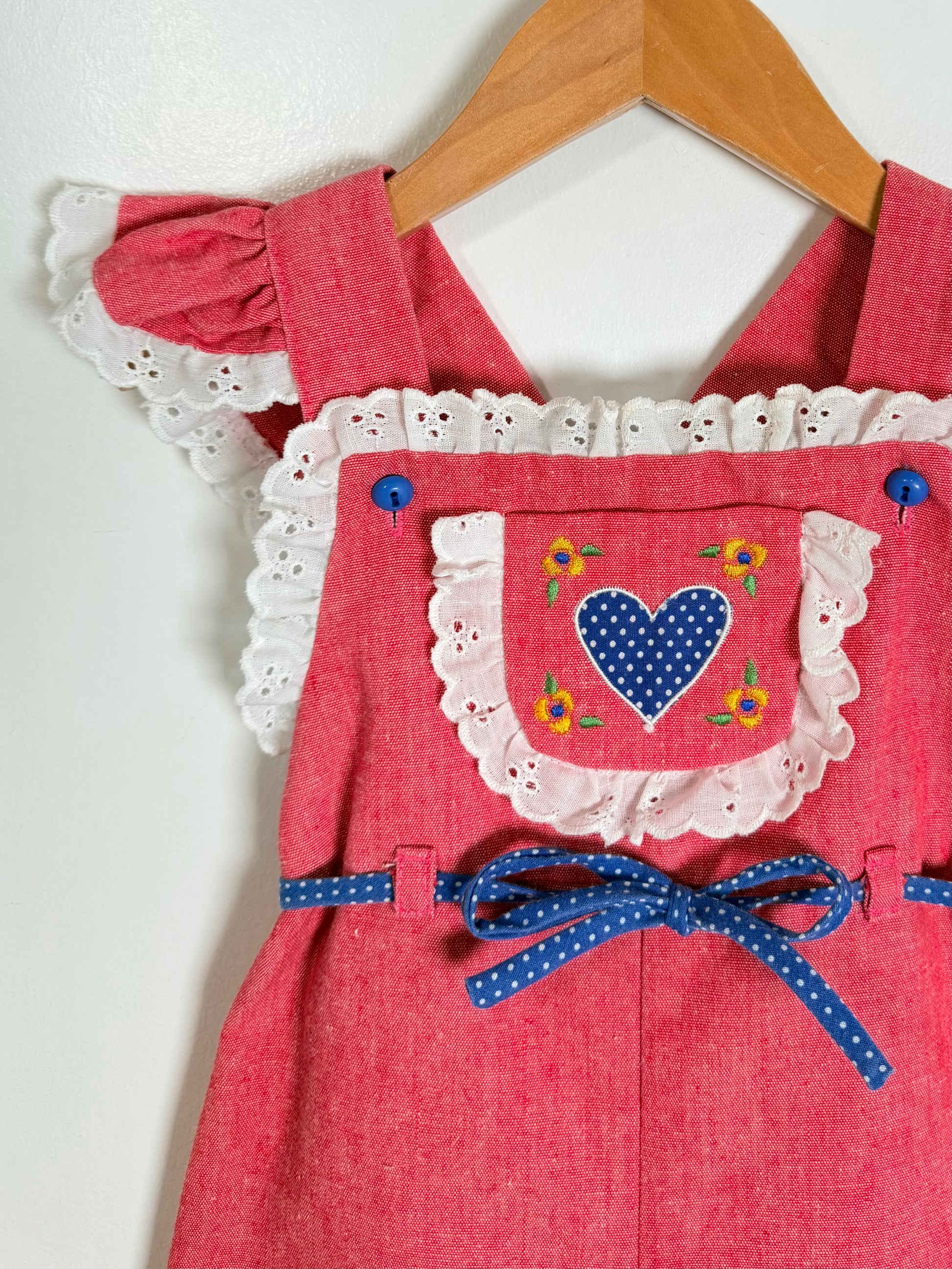 A pink romper with white eyelet lace trim, featuring a front pocket decorated with a blue polka-dot heart, yellow flowers, and a blue polka-dot ribbon bow at the waist, hanging on a wooden hanger.