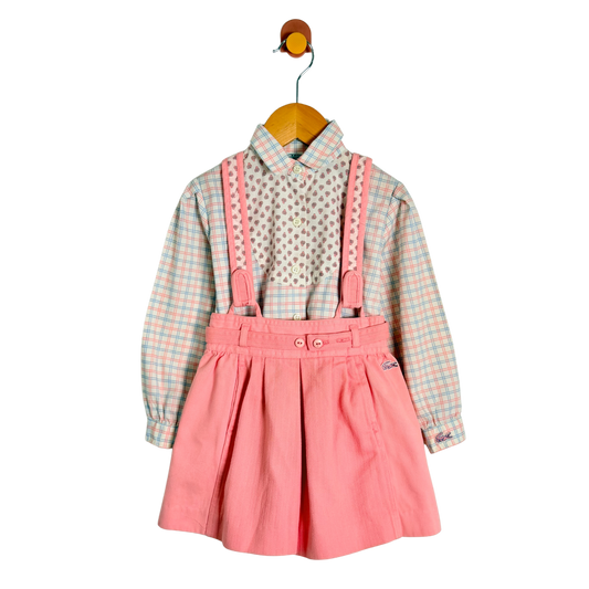 A toddler girl's two-piece outfit hanging on a wooden hanger against a white background. The set includes a long-sleeve button-up shirt with a light pink, green, and white gingham pattern, and a matching pleated pink shorts with pink suspenders that have white lace trim.