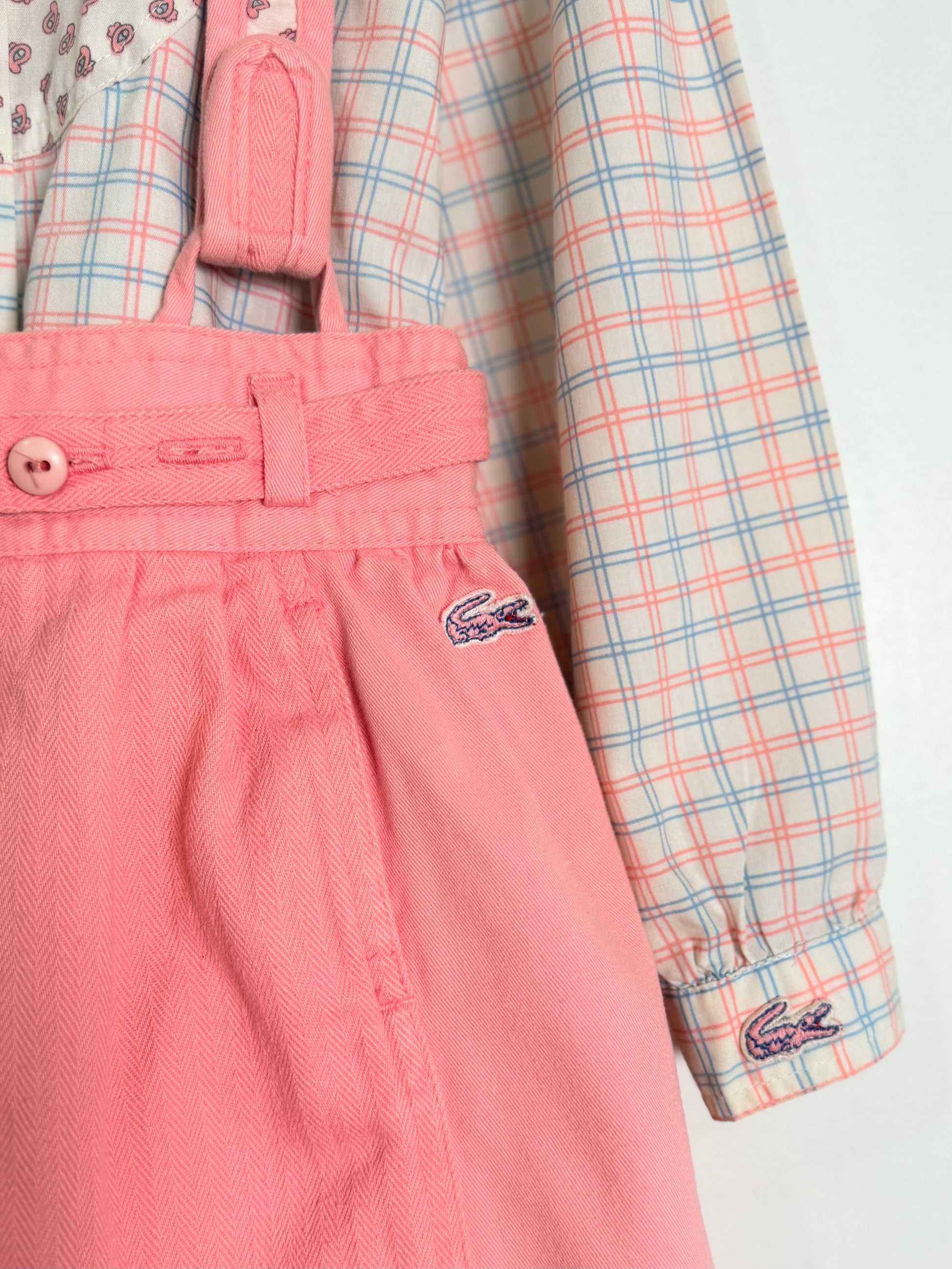 A close-up of a children's outfit featuring pink overalls with a small embroidered Lacoste crocodile logo on the pocket, layered over a pink, blue, and white plaid long-sleeve shirt.