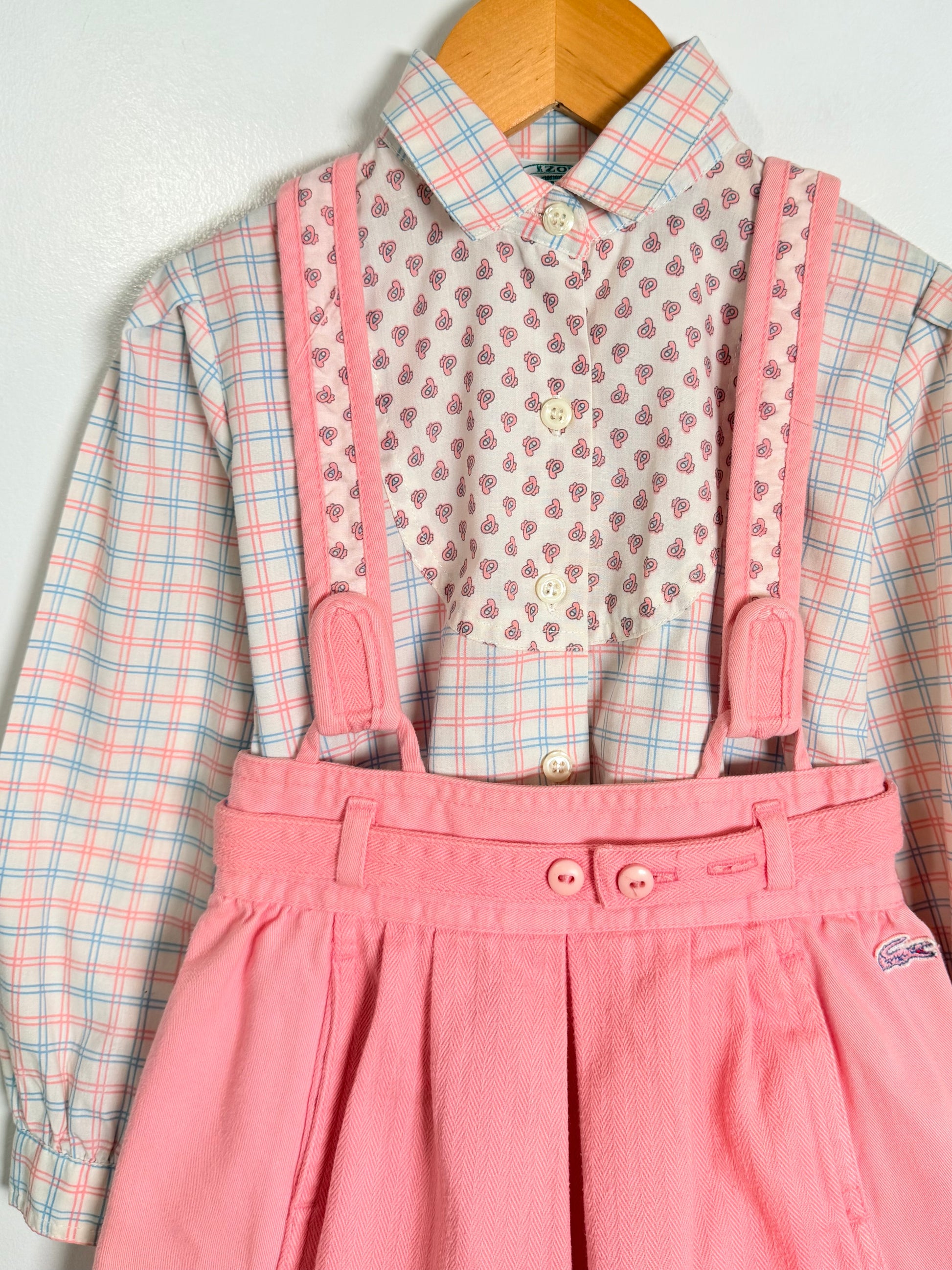 A children's outfit consisting of a pink and blue plaid long-sleeve button-up shirt layered under a pink patterned collared shirt, paired with pink corduroy-style suspenders and a pleated skirt, hanging on a wooden hanger against a light wall.