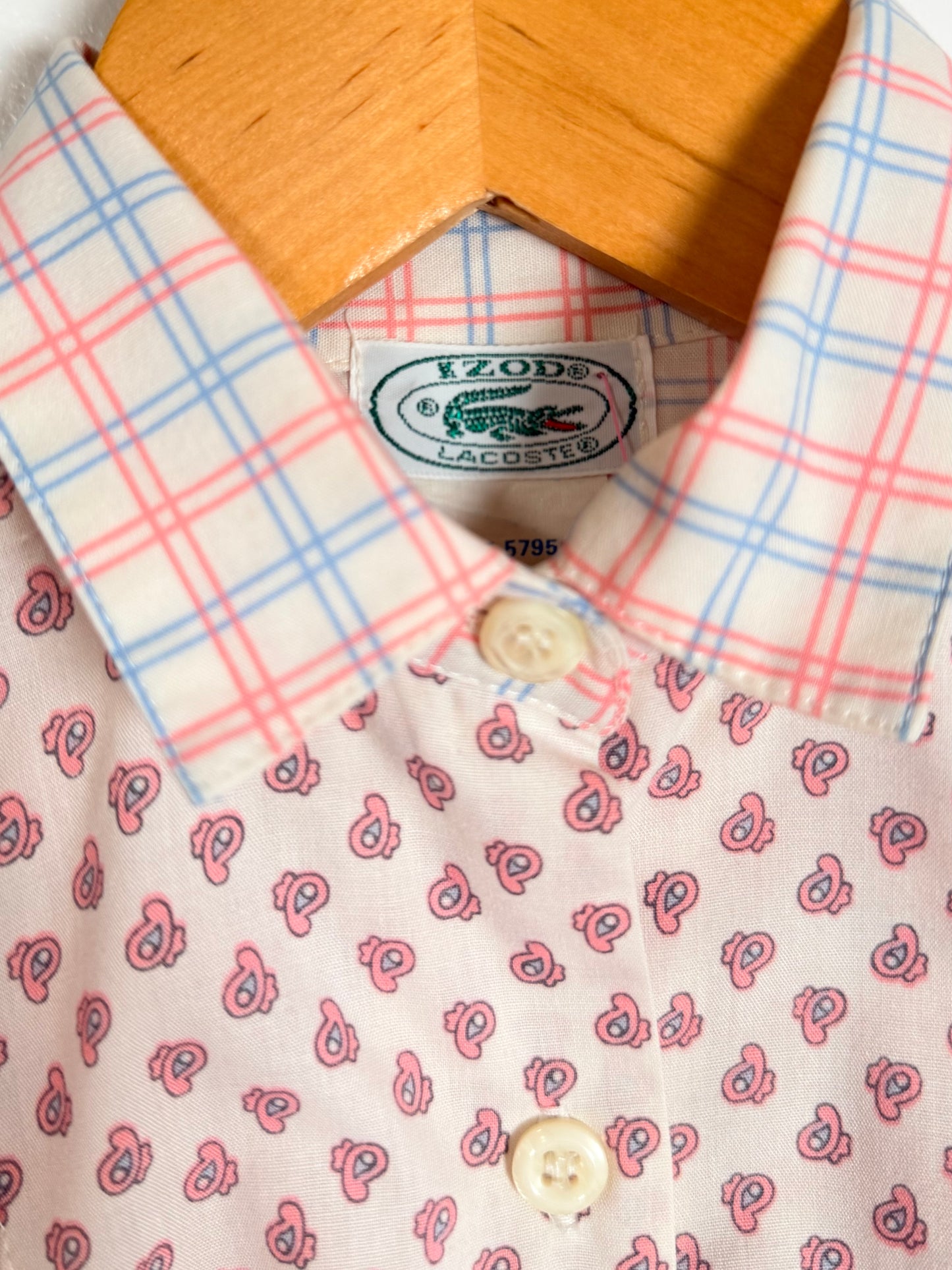 A close-up of a Lacoste button-down shirt on a wooden hanger, featuring a white base with a pink and blue plaid collar and a pink teddy bear pattern on the body.