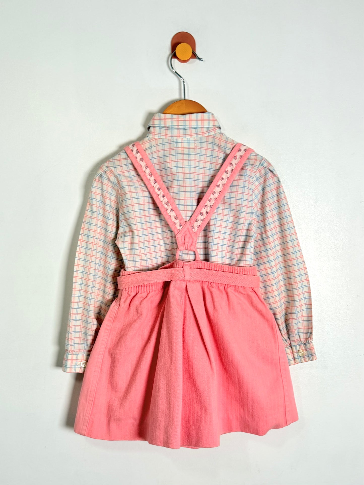 A children's long-sleeve top and skirt set hanging on a wooden hanger against a plain light background. The back view shows a pink and white gingham shirt-style top layered under solid pink shorts with decorative pink trim along the shoulder straps.