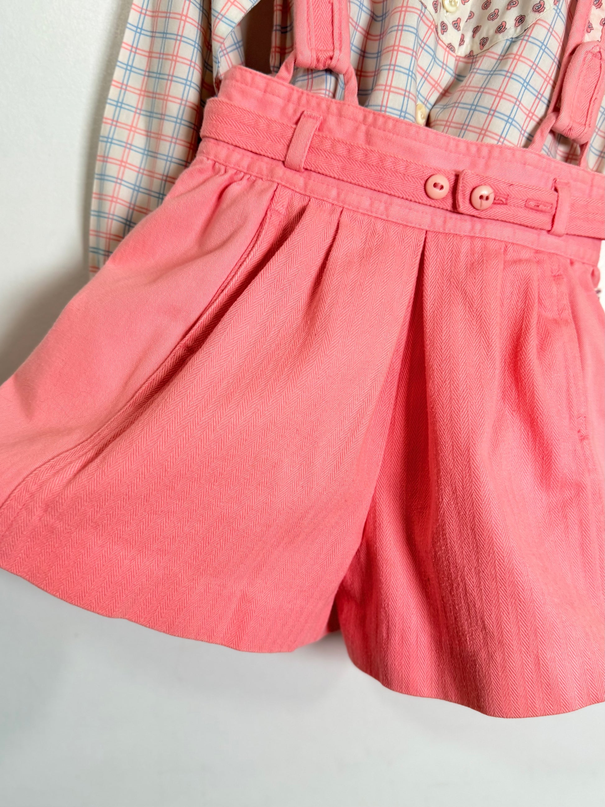 A close-up of a pair of pink pleated shorts with a matching fabric belt and two decorative buttons, shown hanging over a plaid shirt against a plain light background.