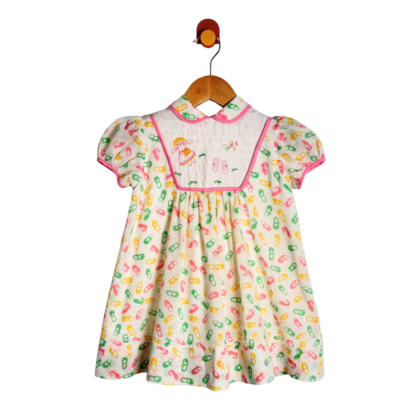 A baby girl's short-sleeve dress hanging on a wooden hanger. The dress has a cream-colored base with a colorful pattern of capsules in pink, green, and yellow, a pink collar, and a smocked yoke featuring embroidered cartoon characters.