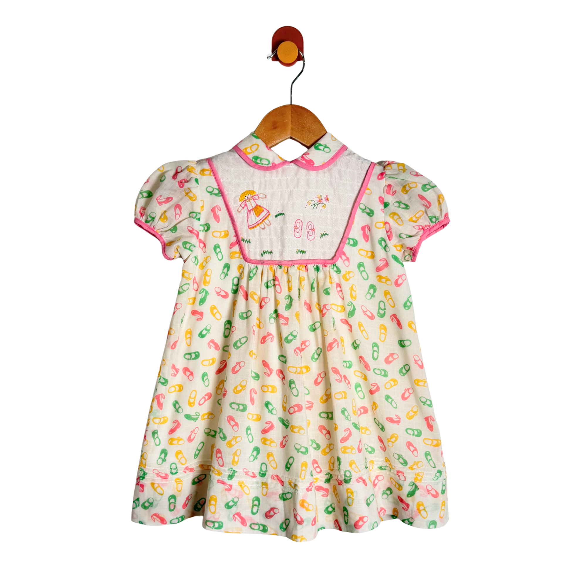 A baby girl's short-sleeve dress hanging on a wooden hanger. The dress has a cream-colored base with a colorful pattern of capsules in pink, green, and yellow, a pink collar, and a smocked yoke featuring embroidered cartoon characters.