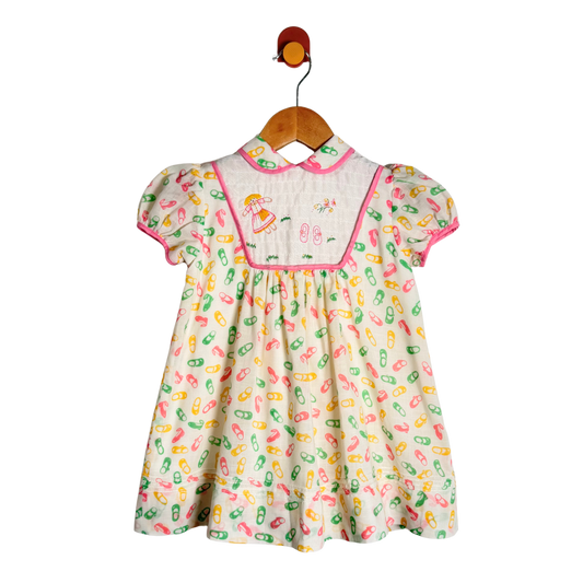 A baby girl's short-sleeve dress hanging on a wooden hanger. The dress has a cream-colored base with a colorful pattern of capsules in pink, green, and yellow, a pink collar, and a smocked yoke featuring embroidered cartoon characters.