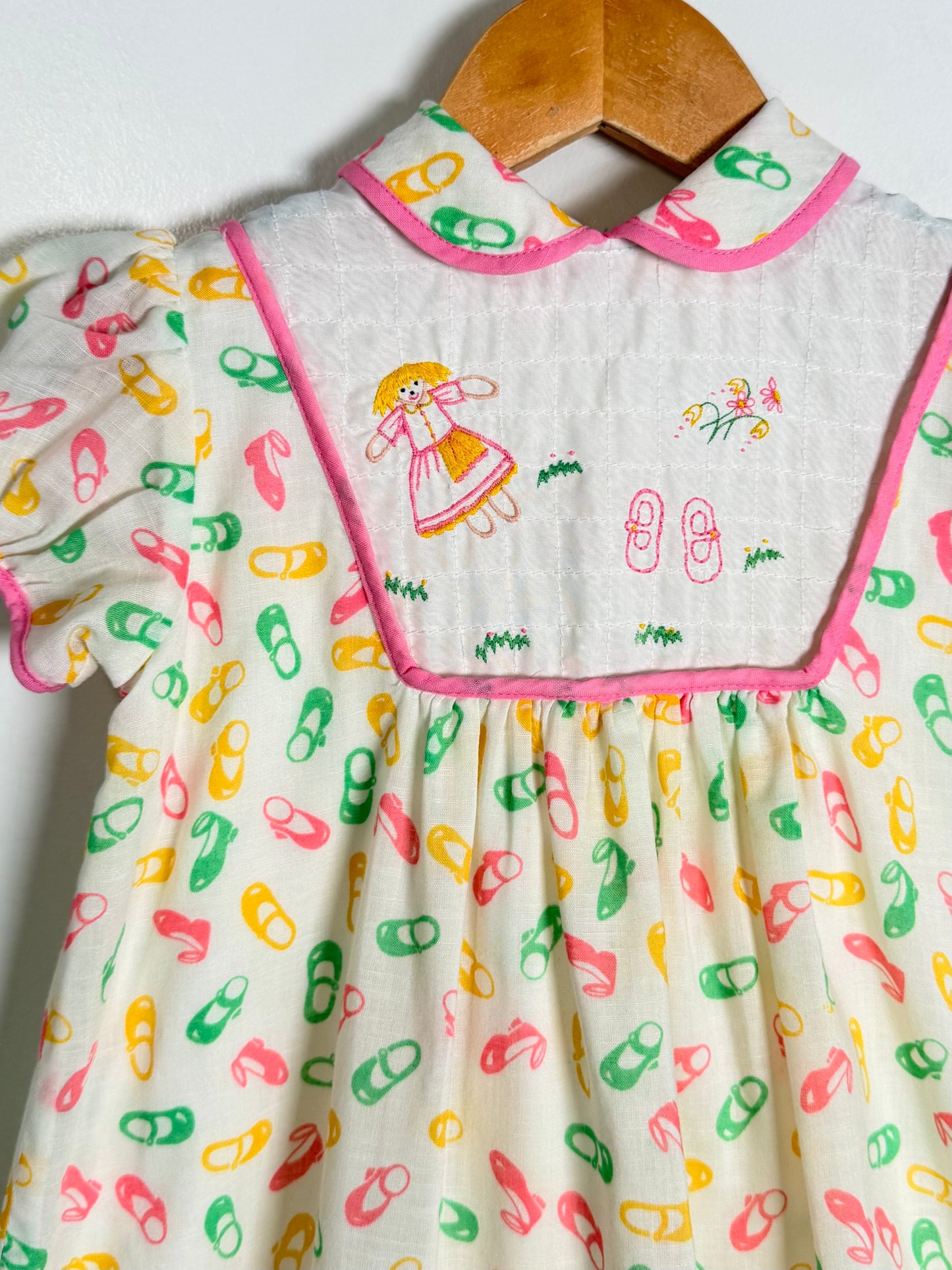 A baby girl's smocked dress with a white bib panel, pink trim, and a colorful print of ballet slippers in pink, green, and yellow. The dress is shown hanging on a wooden hanger against a plain white background.