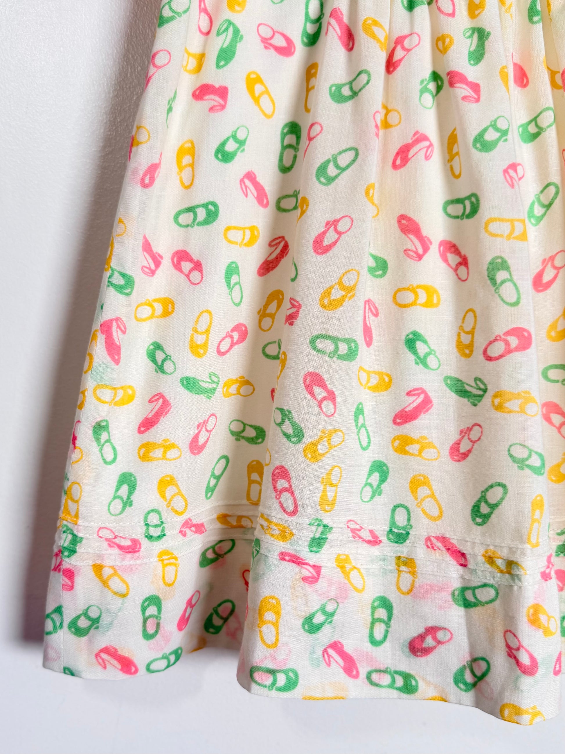 A close-up of a white fabric garment, likely a skirt or dress, featuring an all-over print of colorful shoes in pink, green, and yellow.