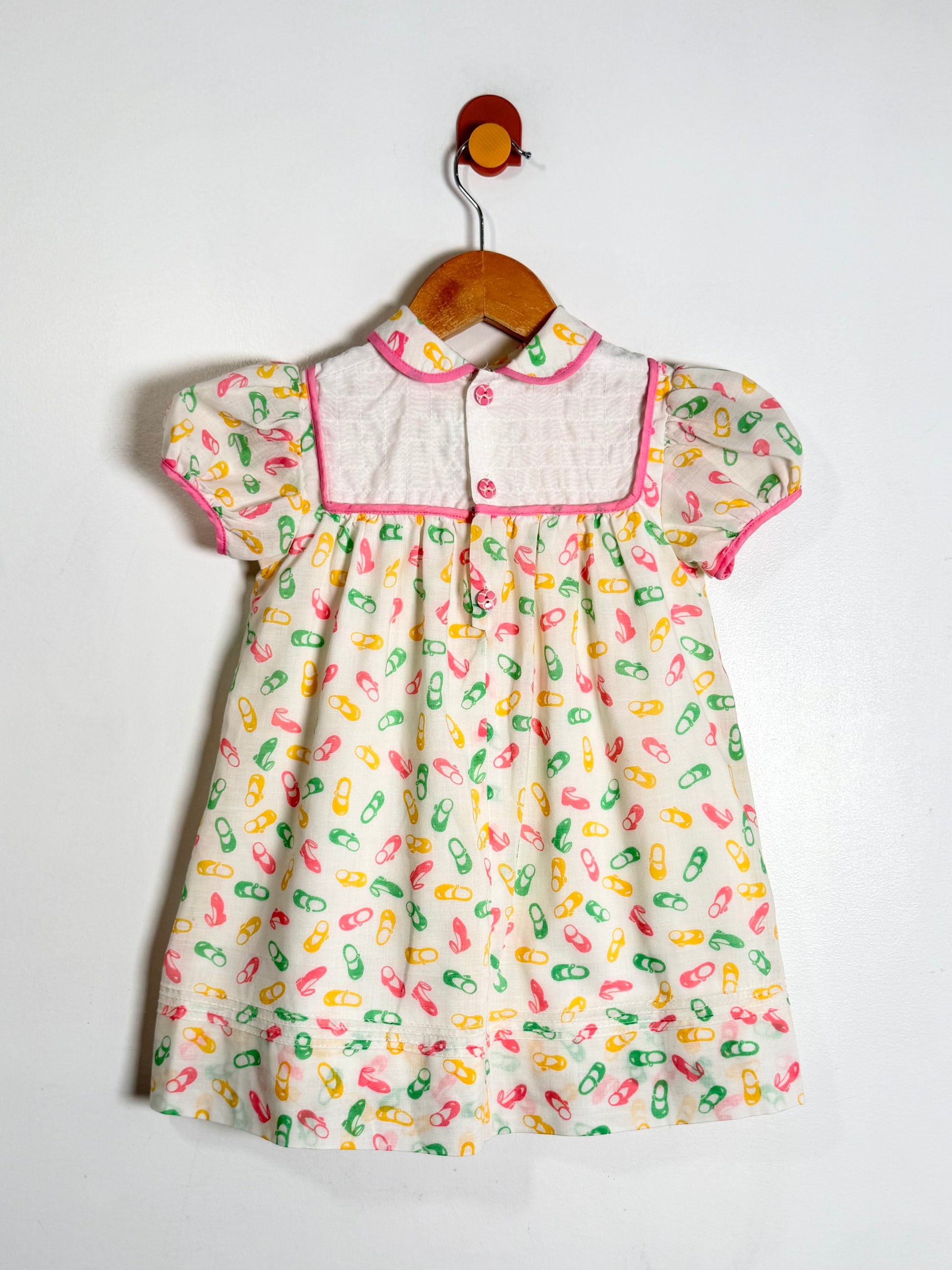 A baby girl's short-sleeve dress hanging on a wooden hanger against a plain white wall. The dress has a white collar with pink piping and buttons, and a cream-colored body covered in a colorful print of shoes in pink, green, and yellow.
