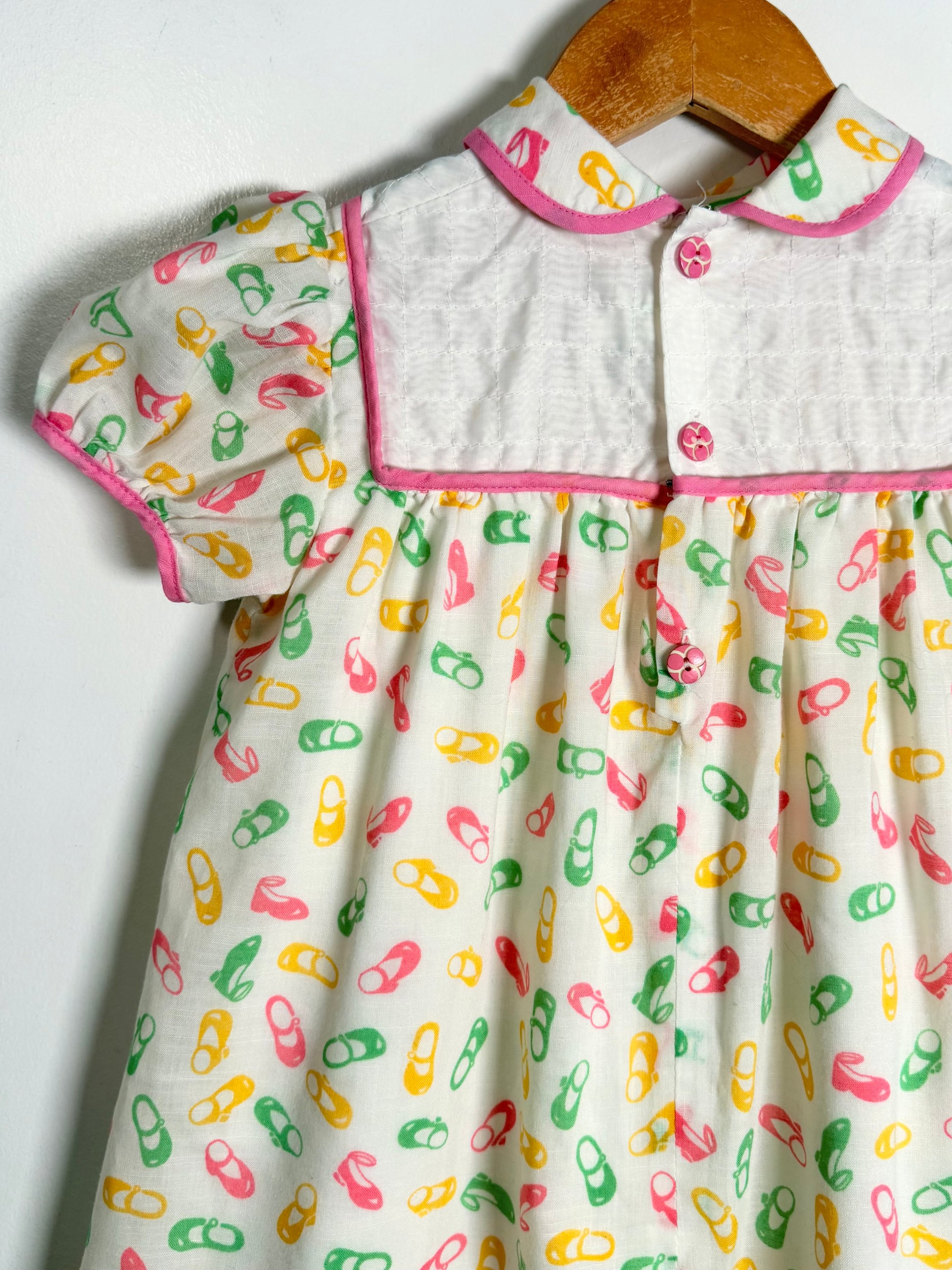 A short-sleeve baby dress hanging on a wooden hanger against a plain white wall. The dress has a white collar and yoke with pink trim, and a white skirt covered in a colorful print of pink, green, and yellow shoes.
