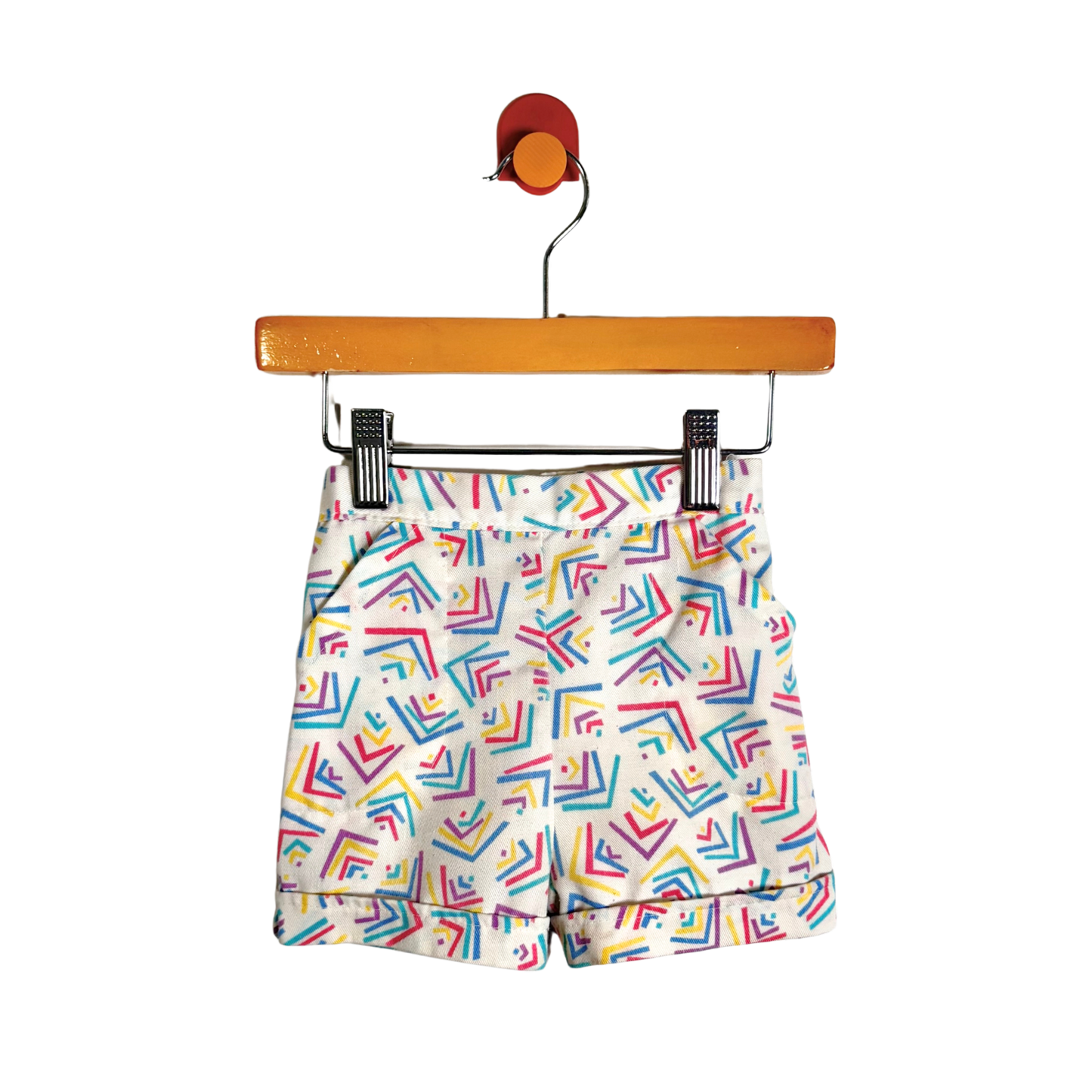 White shorts with a colorful geometric pattern of intersecting lines in red, blue, yellow, and green, hanging from a wooden hanger with black clips against a white background.