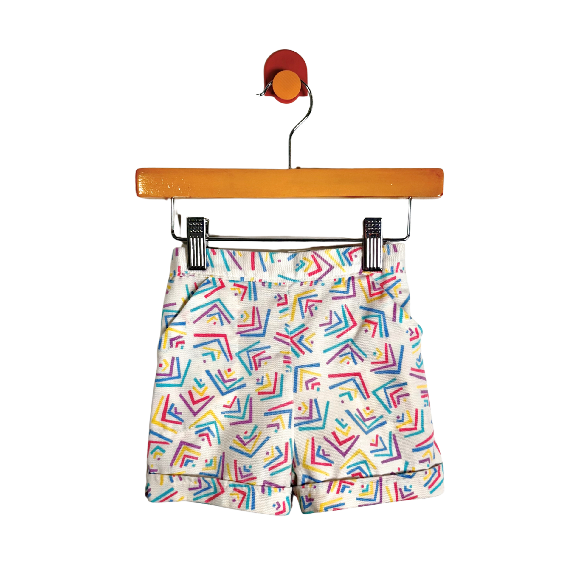 White shorts with a colorful geometric pattern of intersecting lines in red, blue, yellow, and green, hanging from a wooden hanger with black clips against a white background.