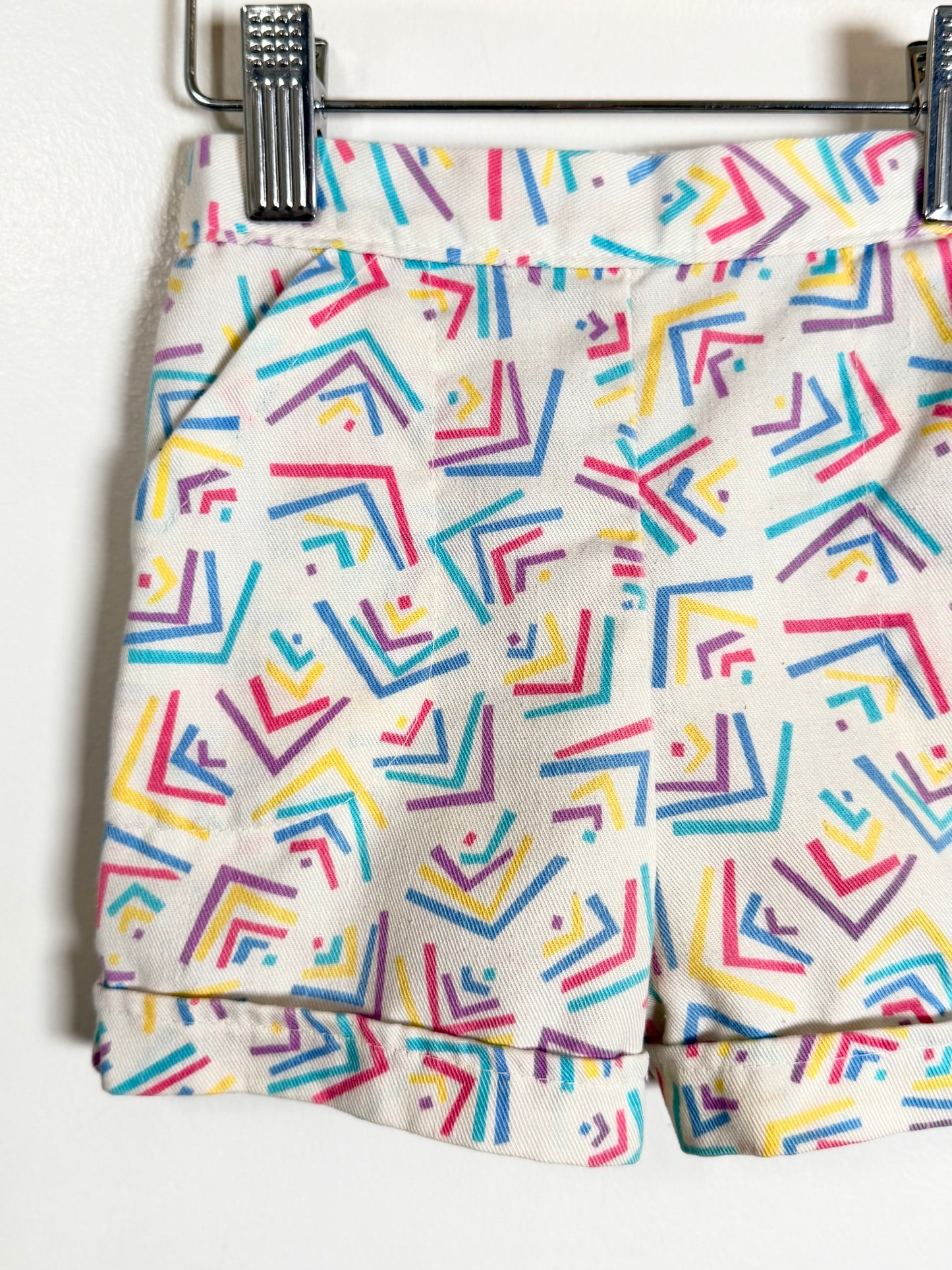 A close-up of a pair of shorts hanging from a metal hanger, featuring a colorful geometric pattern of abstract lines in blue, pink, yellow, and purple on a cream-colored background.