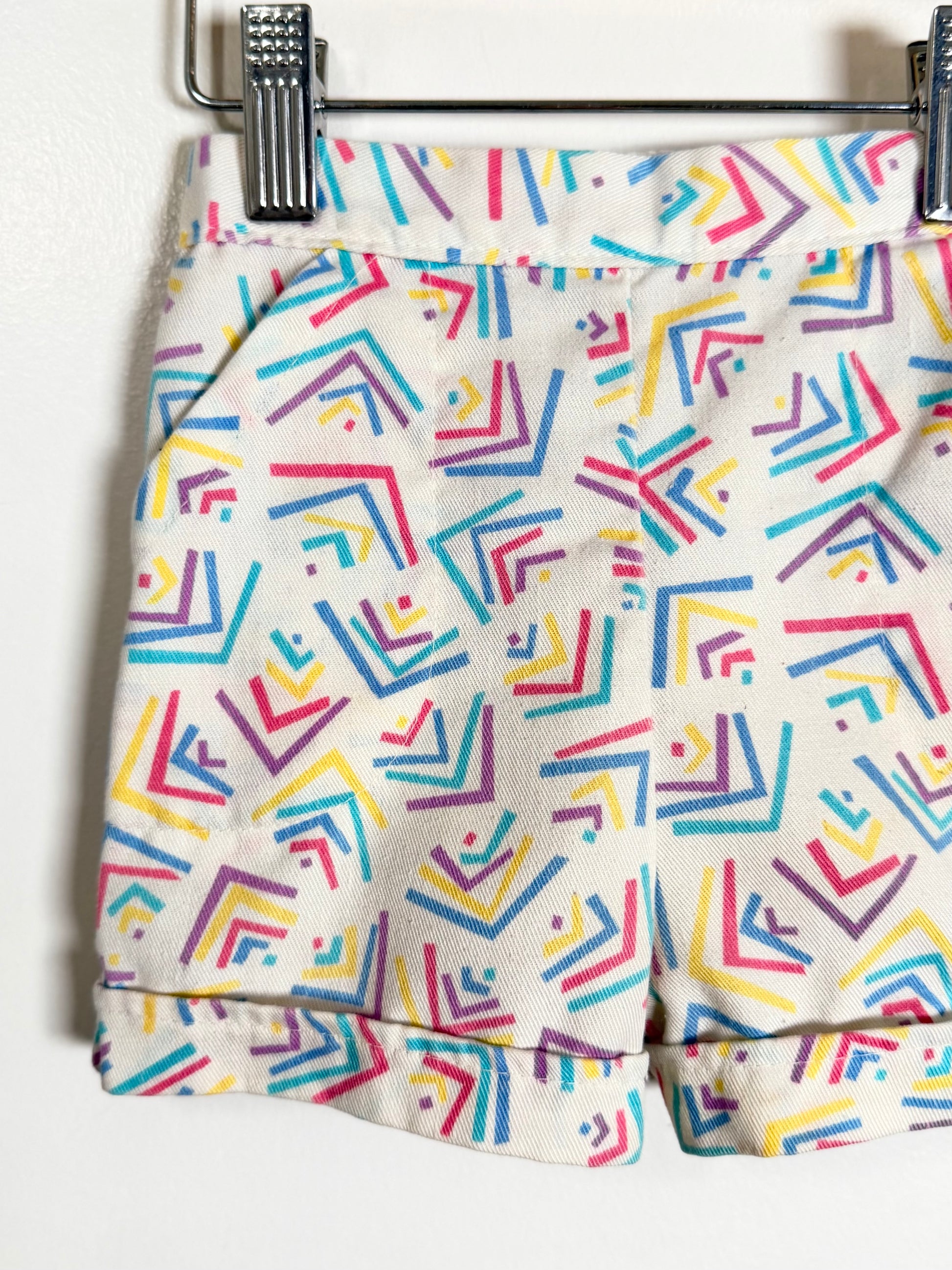 A close-up of a pair of shorts hanging from a metal hanger, featuring a colorful geometric pattern of abstract lines in blue, pink, yellow, and purple on a cream-colored background.