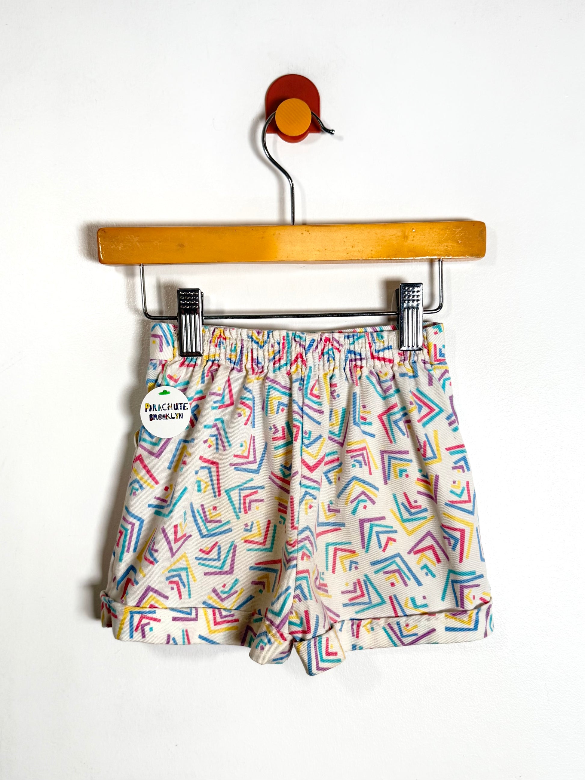 A pair of children's shorts with a colorful geometric pattern on a cream background, hanging from a wooden hanger against a plain white wall.