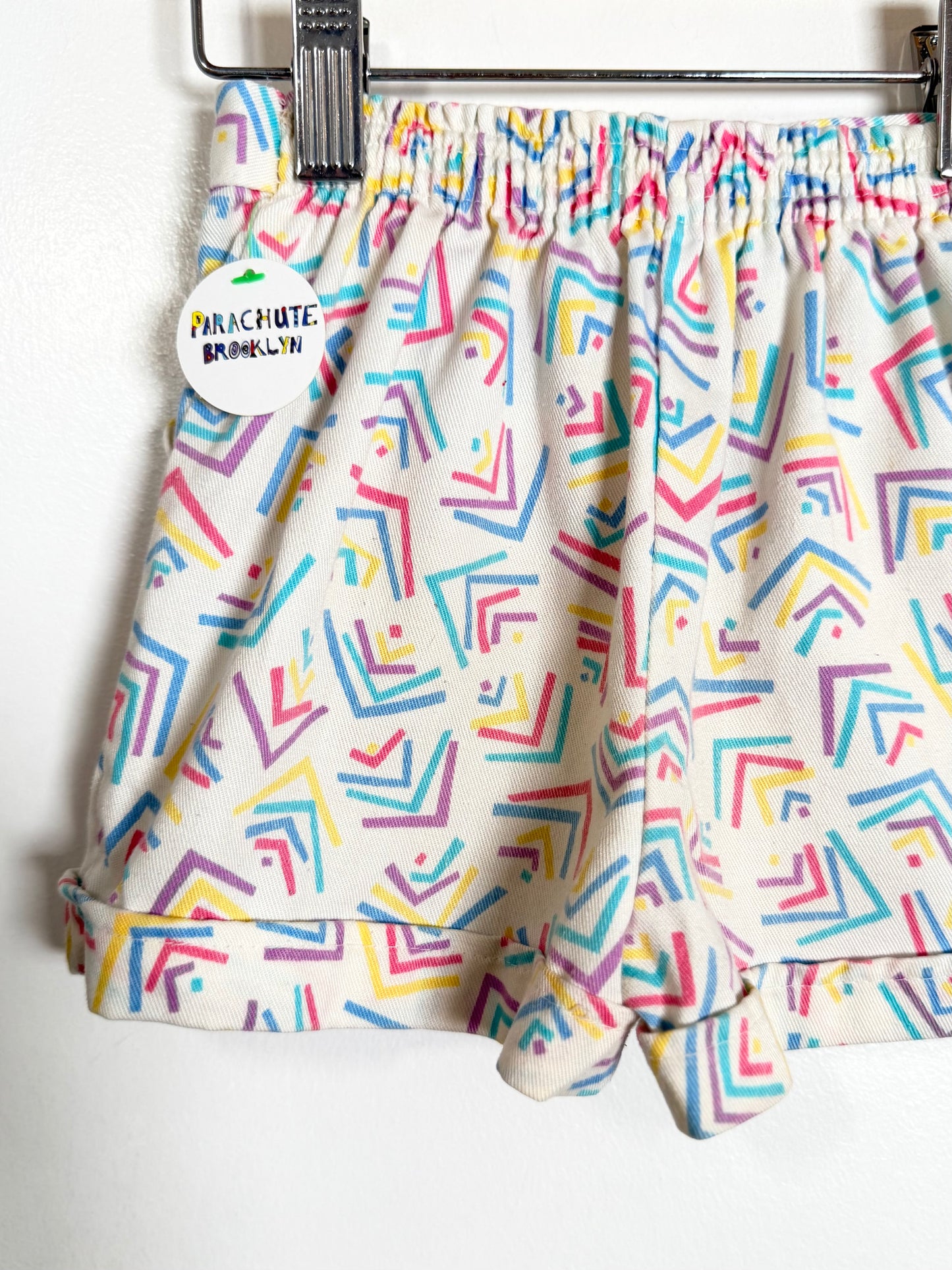 A pair of children's shorts with a colorful geometric pattern on a cream background, hanging from a metal hanger with a brand tag 