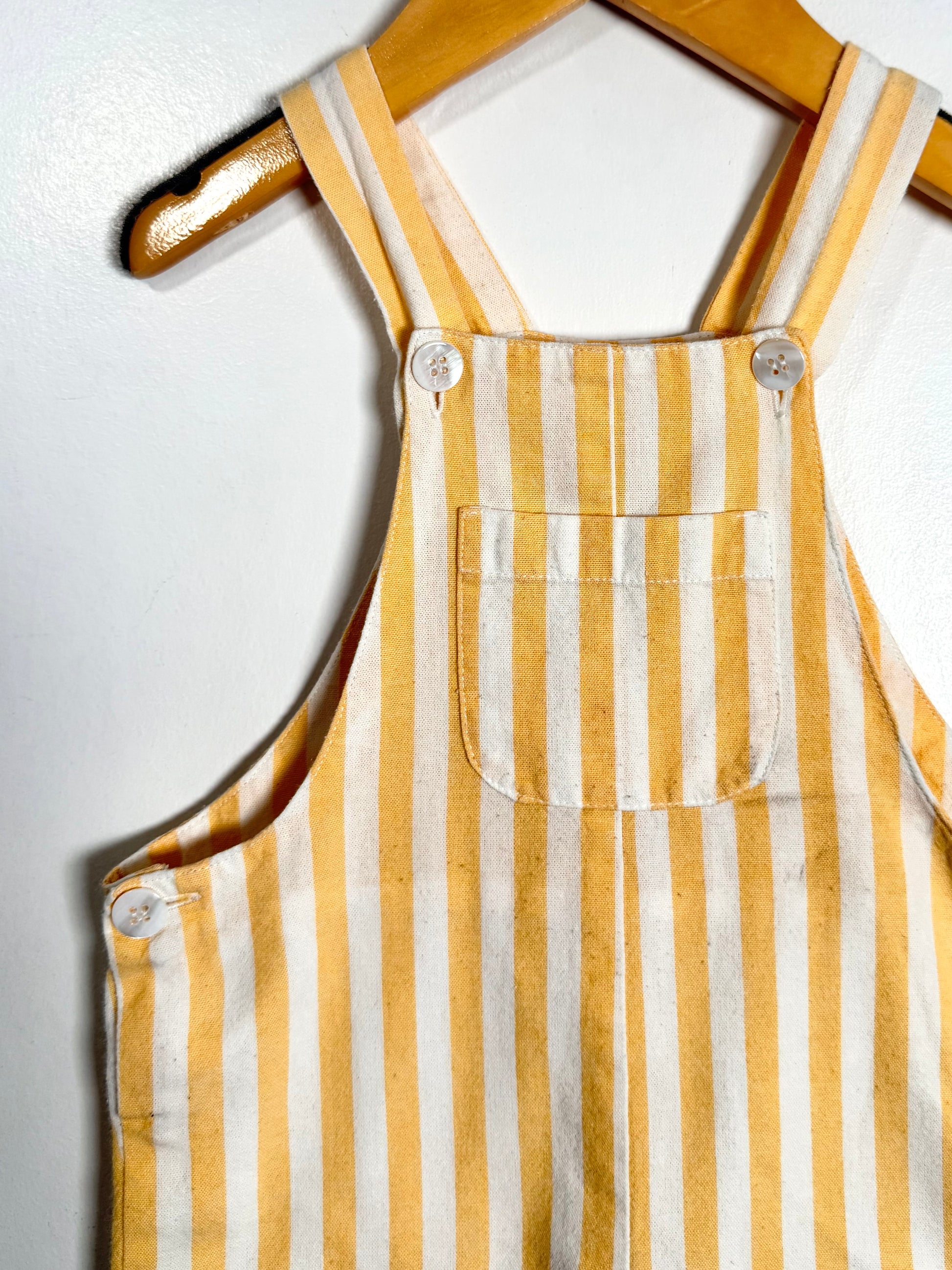 Yellow and white striped bib overalls hanging on a wooden hanger against a plain white wall.
