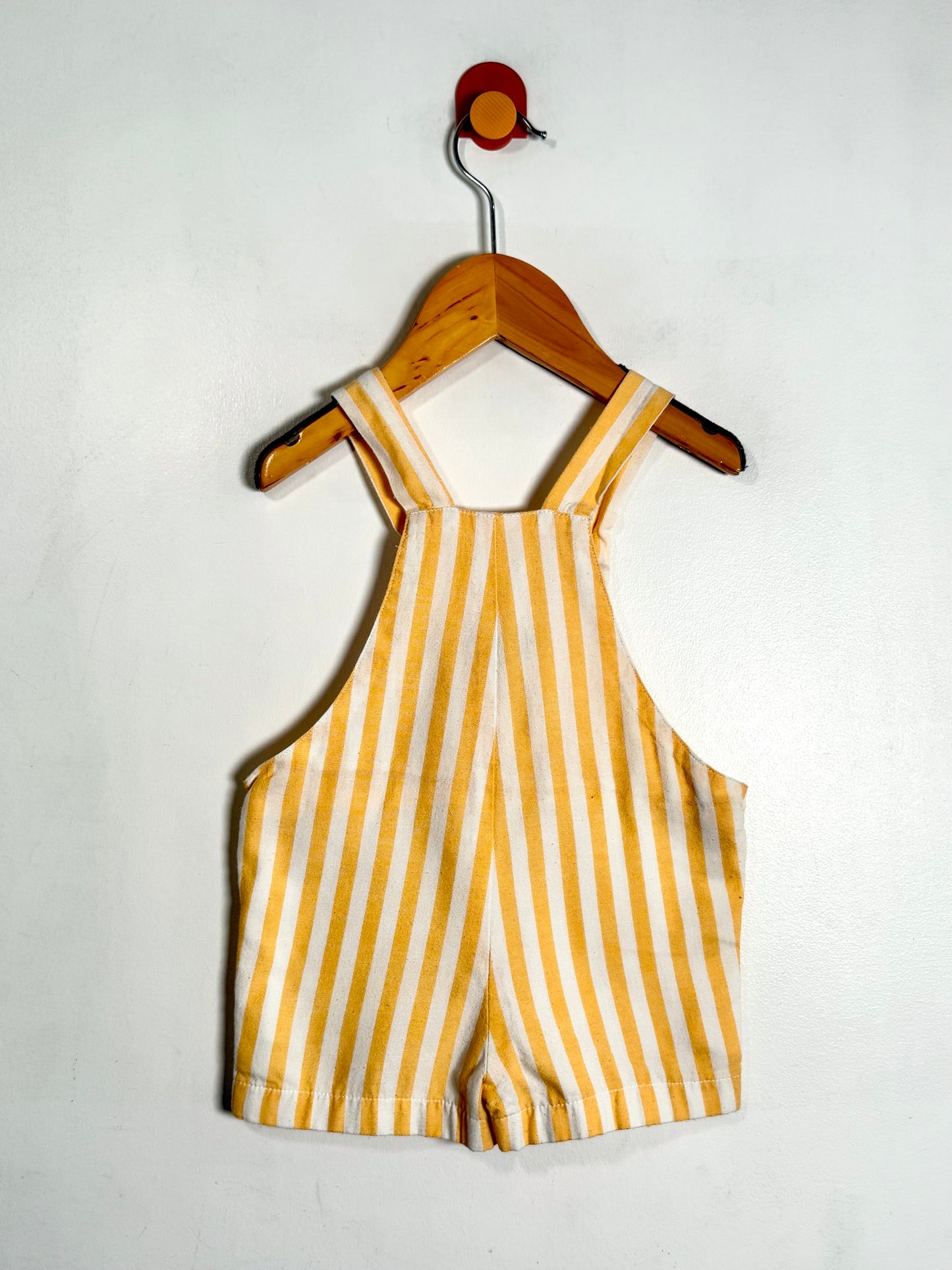 A baby romper with vertical yellow and white stripes, hanging on a wooden hanger with a red hook against a plain white wall.