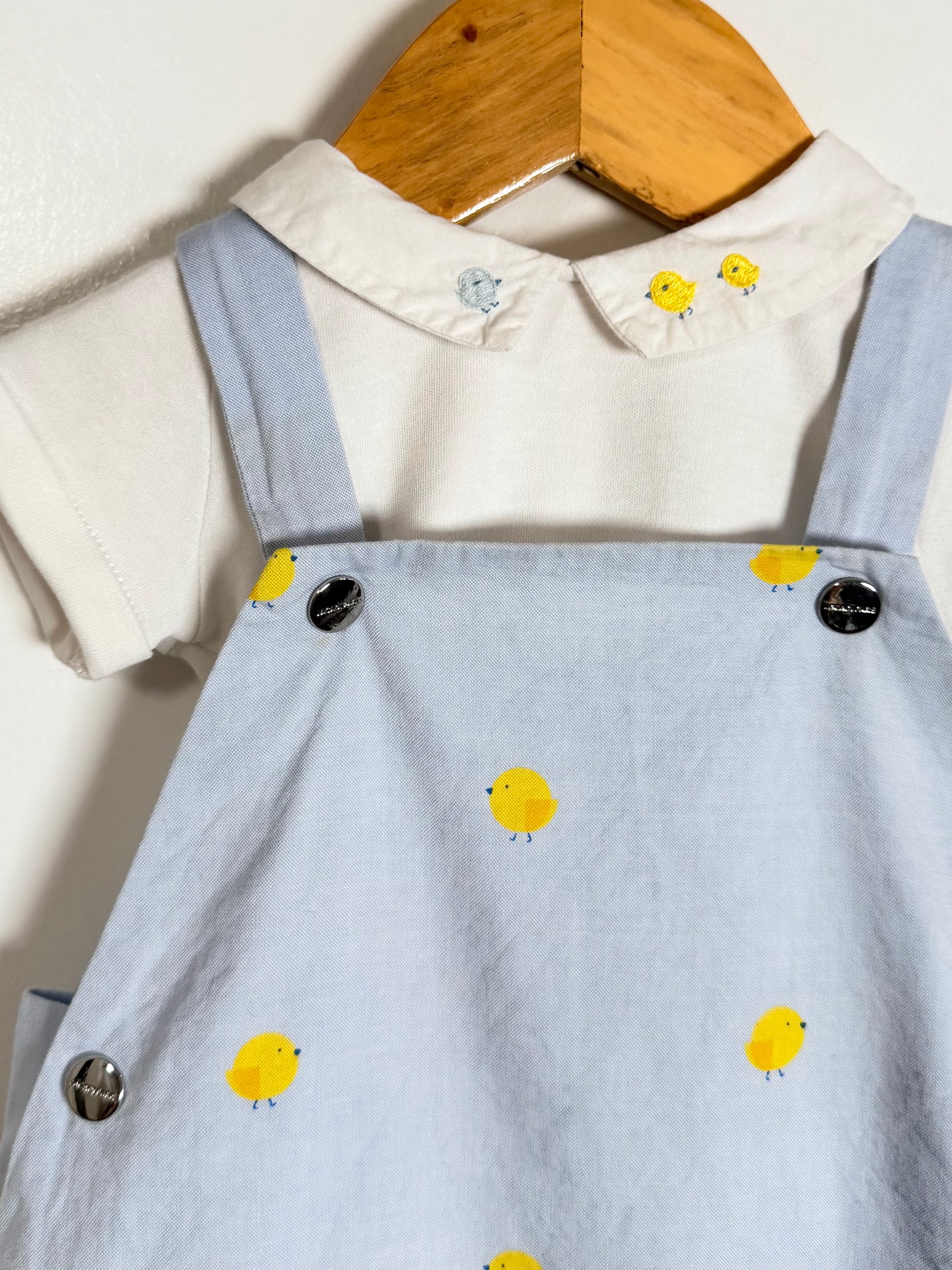 A baby short-sleeve outfit featuring a white collared shirt with yellow chick details on the collar, layered under a light blue denim-style overall dress with yellow chick prints and silver buttons.