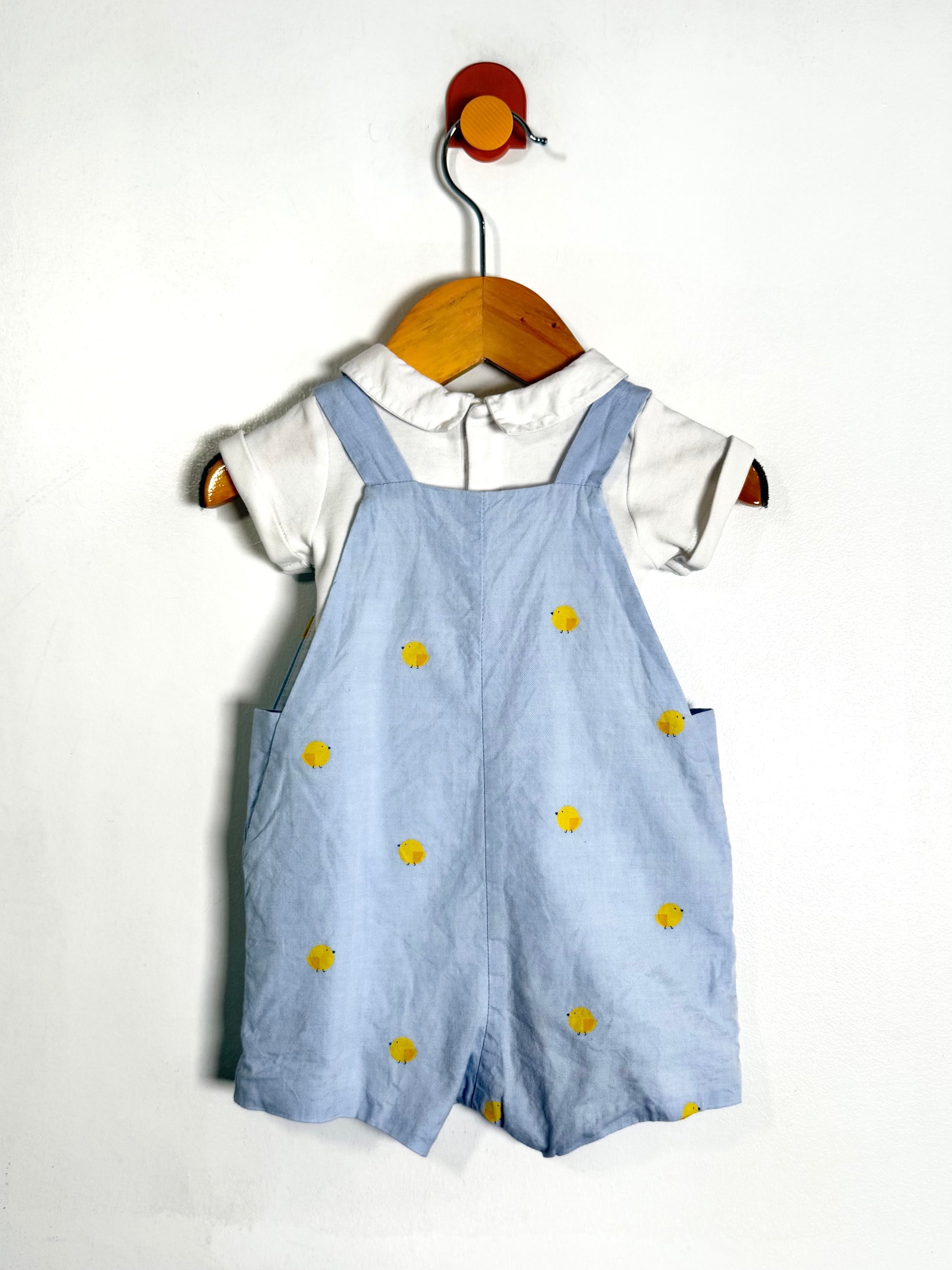 A baby romper set consisting of light blue denim-style overall shorts with yellow chicks, worn over a white short-sleeve collared shirt, hanging on a wooden hanger against a plain white wall.