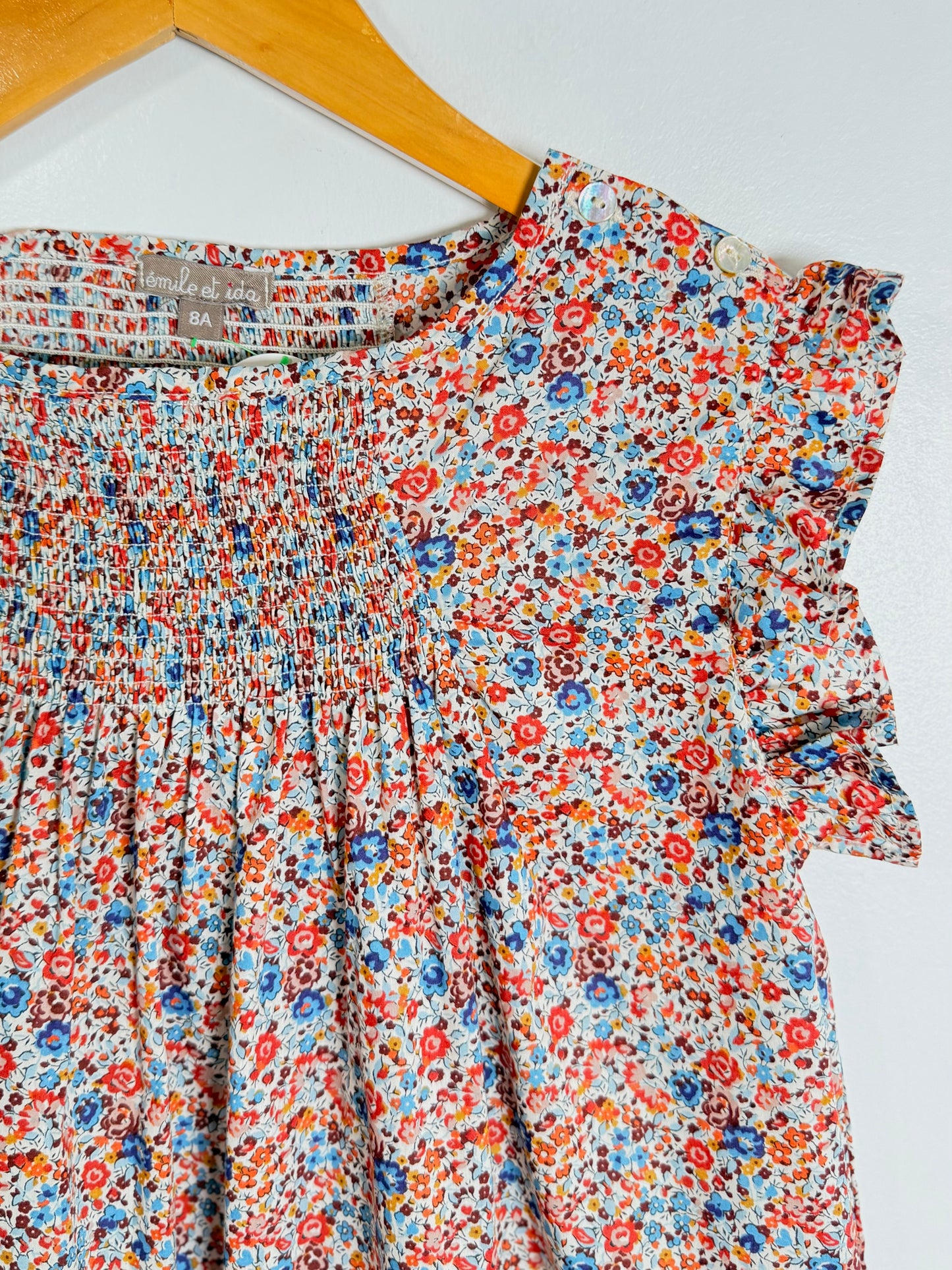 A close-up of a floral print dress hanging on a wooden hanger against a white wall. The dress features a dense multicolor floral pattern with red, blue, orange, and yellow flowers on a light background, with a smocked bodice and ruffled sleeves.