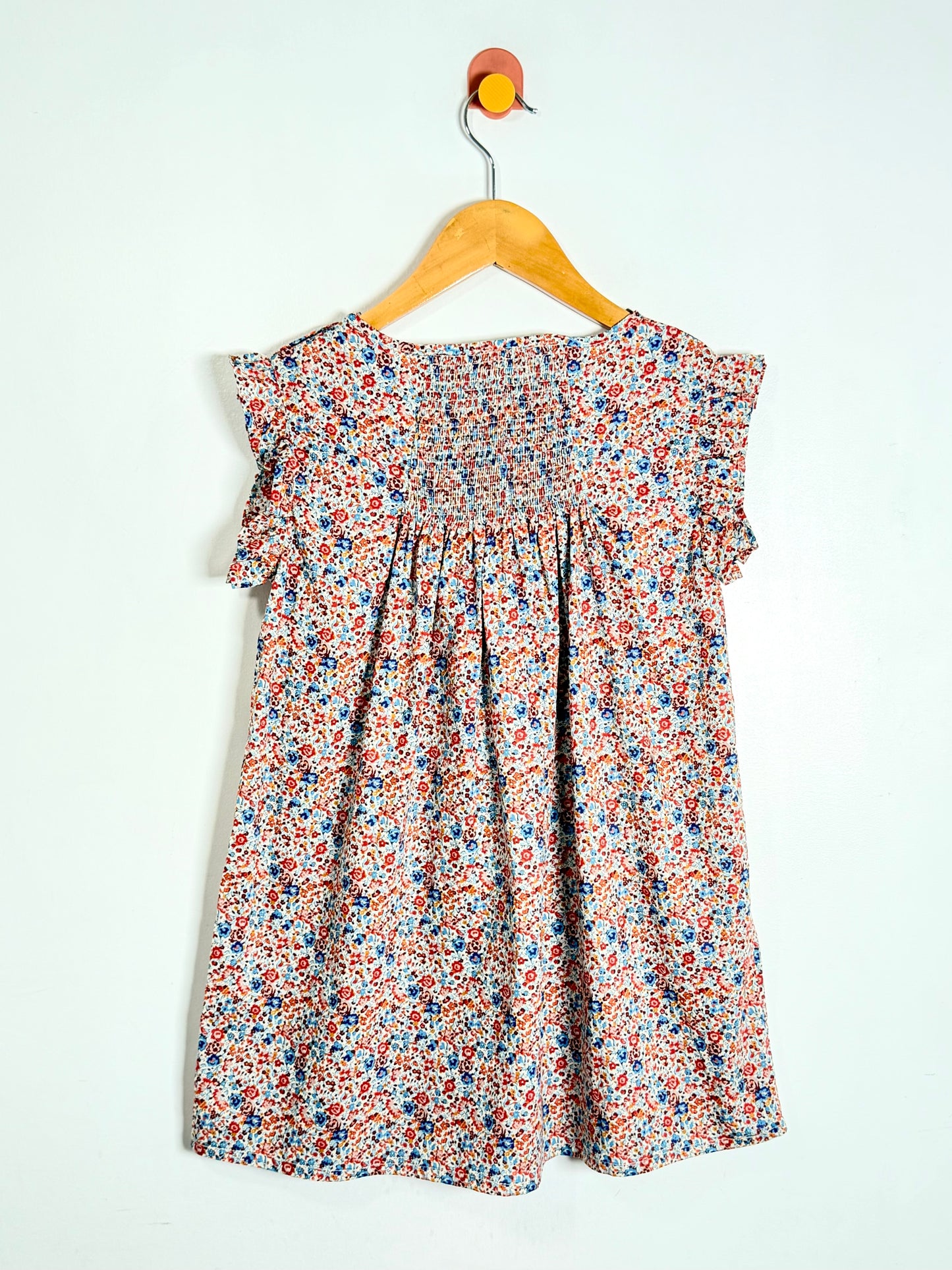 A sleeveless children's dress with a smocked bodice and ruffled shoulders, featuring a dense multicolor floral print in red, blue, and orange on a light background, hanging on a wooden hanger against a plain white wall.