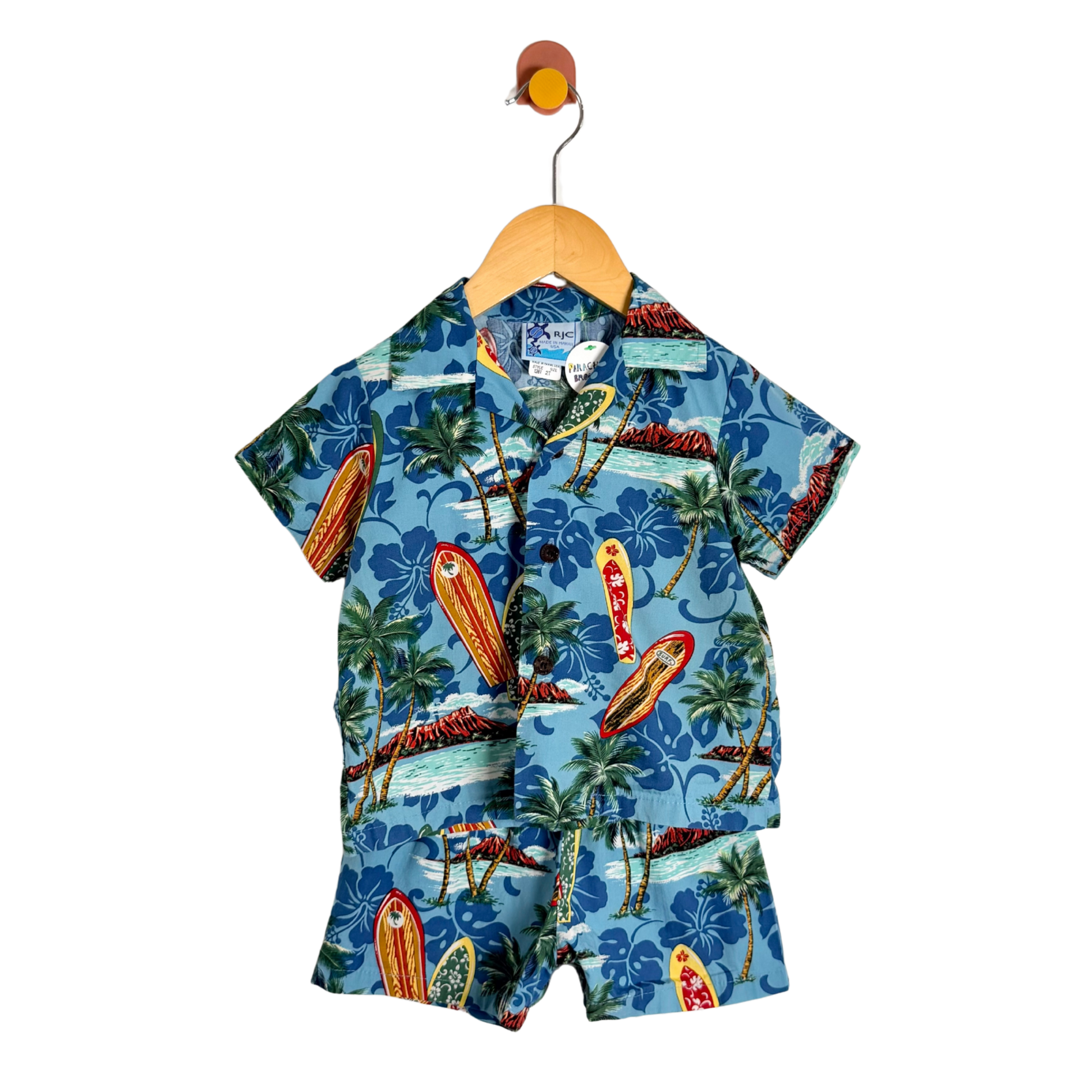 A children's short-sleeve button-up shirt and matching shorts set in a blue tropical surfboard and palm tree print, hanging on a wooden hanger against a white background.