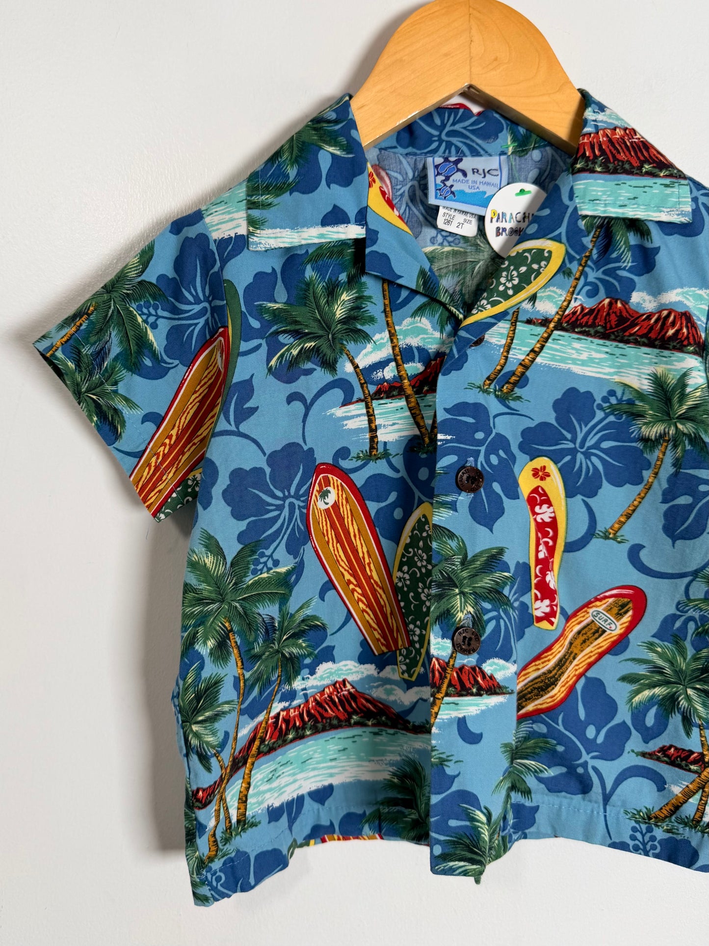 A short-sleeve button-up shirt with a blue tropical beach print, hanging on a wooden hanger against a plain white background. The print features palm trees, surfboards, and hibiscus flowers.
