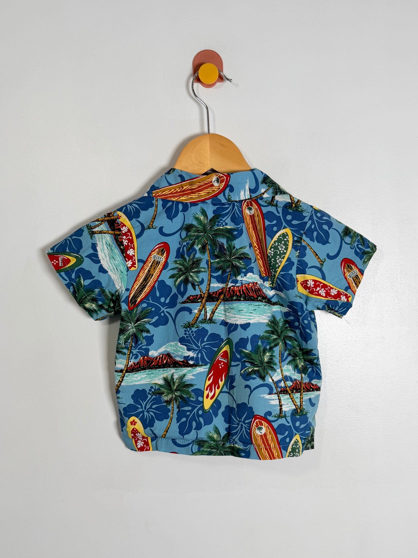 A short-sleeve children's button-up shirt with a tropical beach print, hanging on a wooden hanger against a plain white wall. The shirt features a blue background with surfboards, palm trees, and islands.