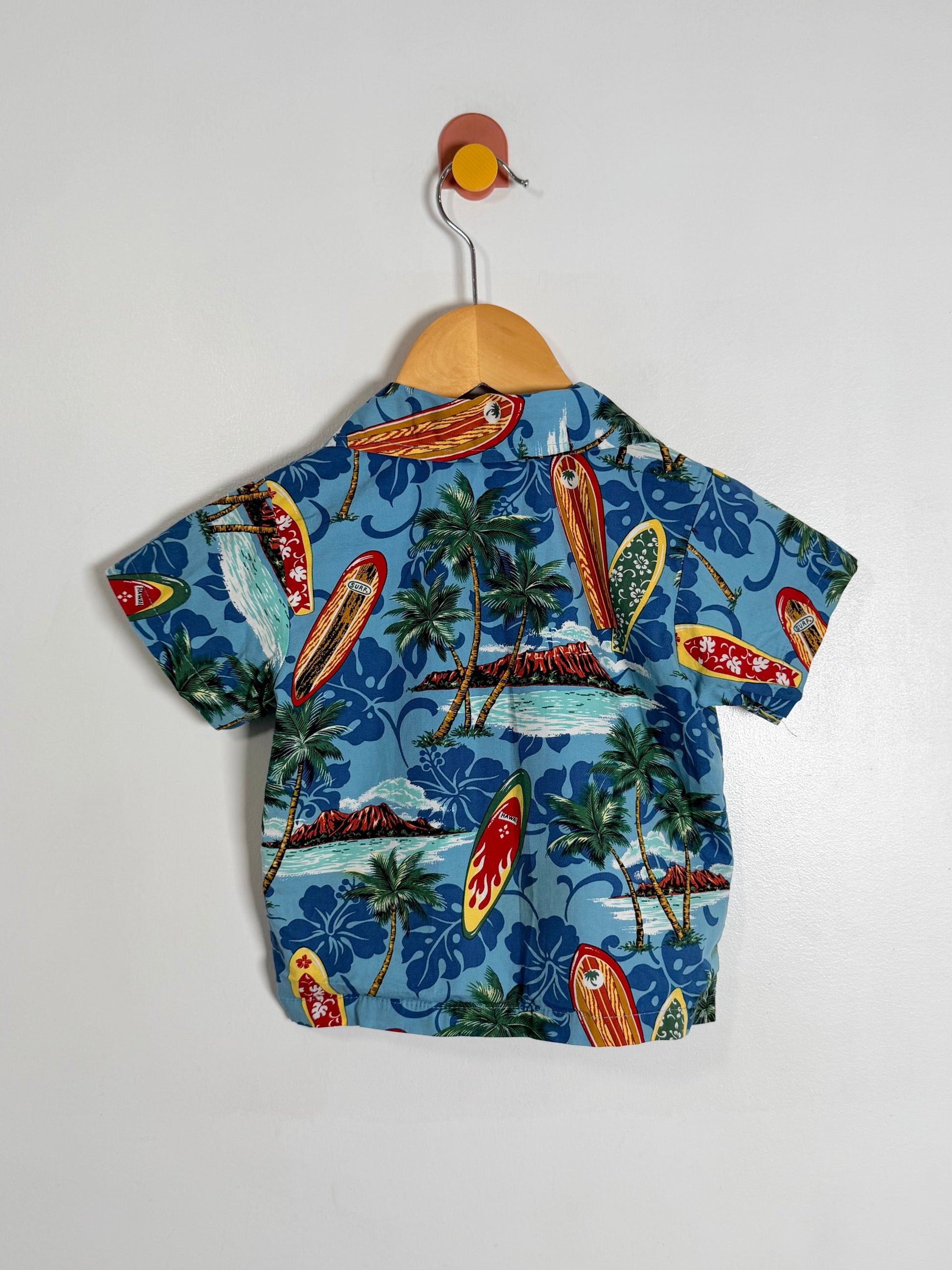 A short-sleeve children's button-up shirt with a tropical beach print, hanging on a wooden hanger against a plain white wall. The shirt features a blue background with surfboards, palm trees, and islands.