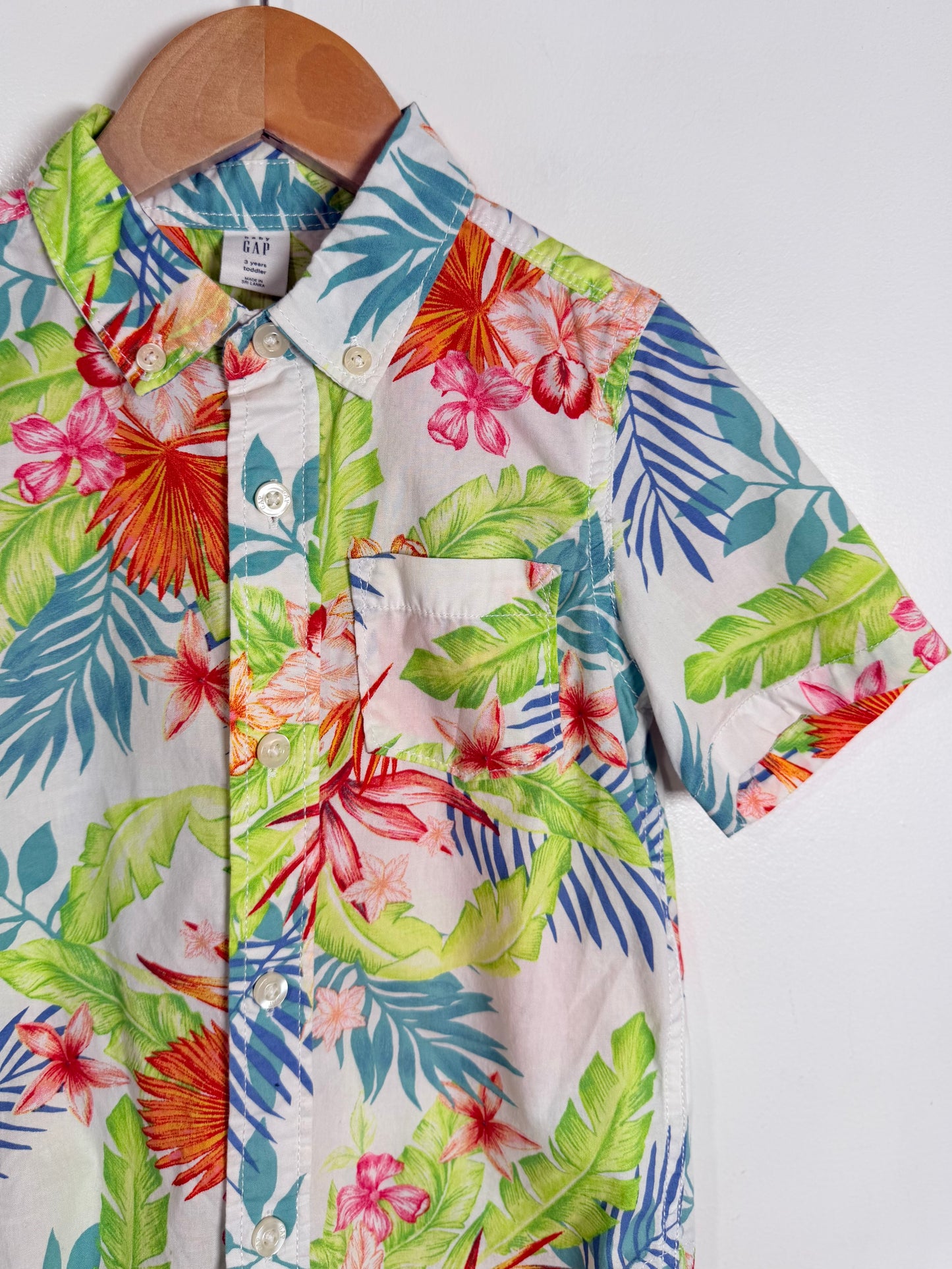 A short-sleeve button-up shirt with a vibrant tropical floral and leaf print, hanging on a wooden hanger against a plain light gray background.