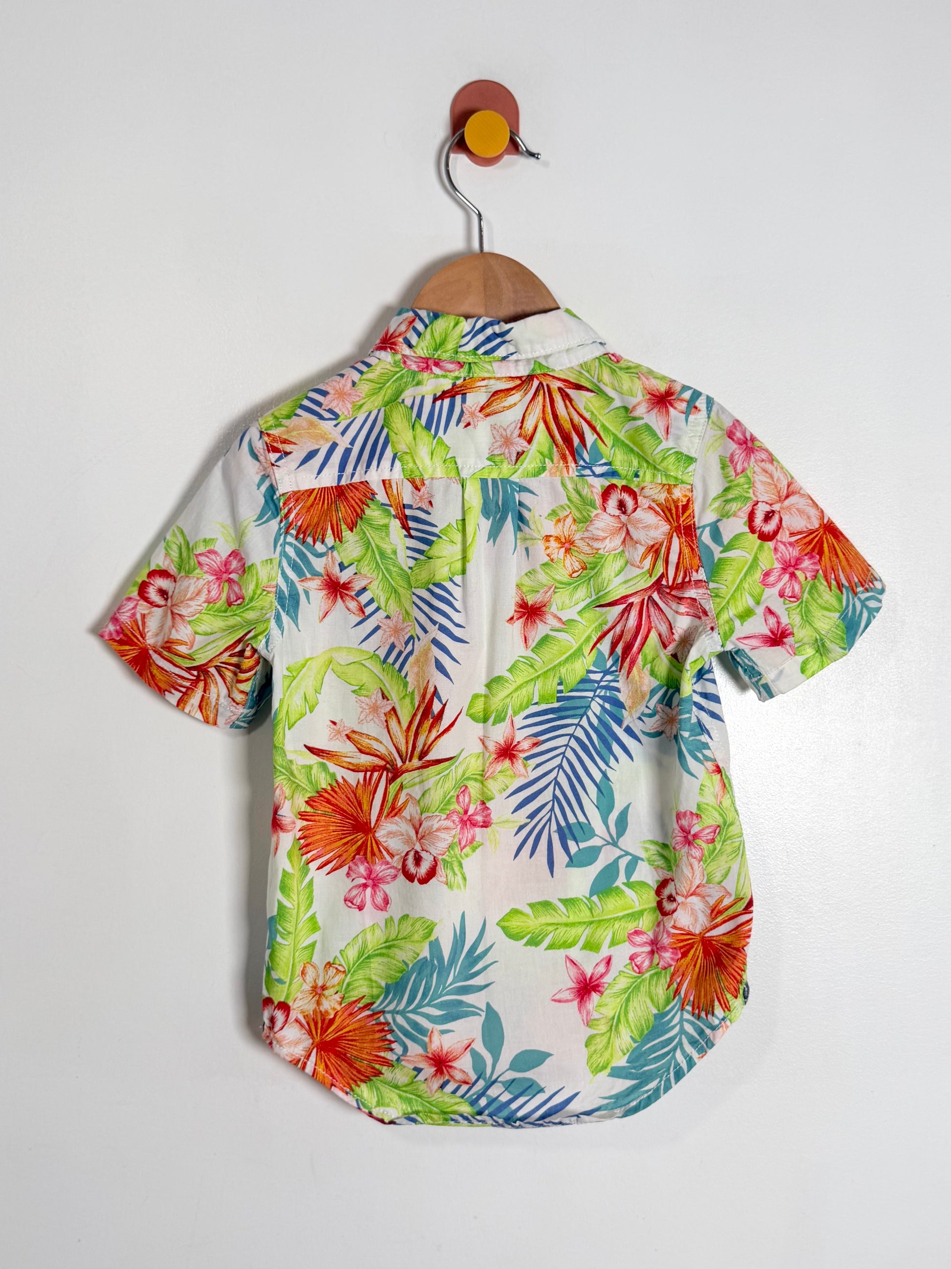 A short-sleeve button-up shirt with a vibrant tropical floral and leaf print, hanging on a wooden hanger against a plain white wall.