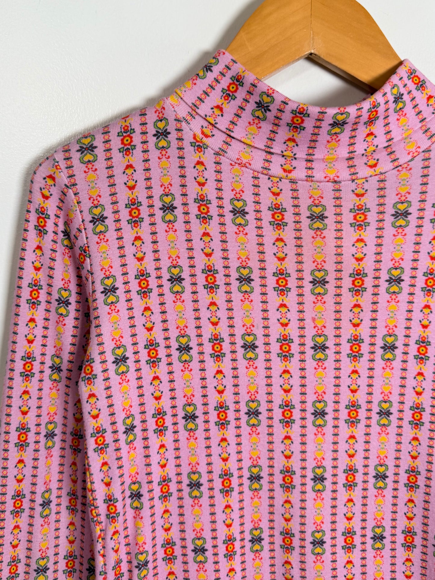 A pink long-sleeve mock neck top with a colorful vertical pattern of small flowers and geometric shapes, hanging on a wooden hanger against a white wall.