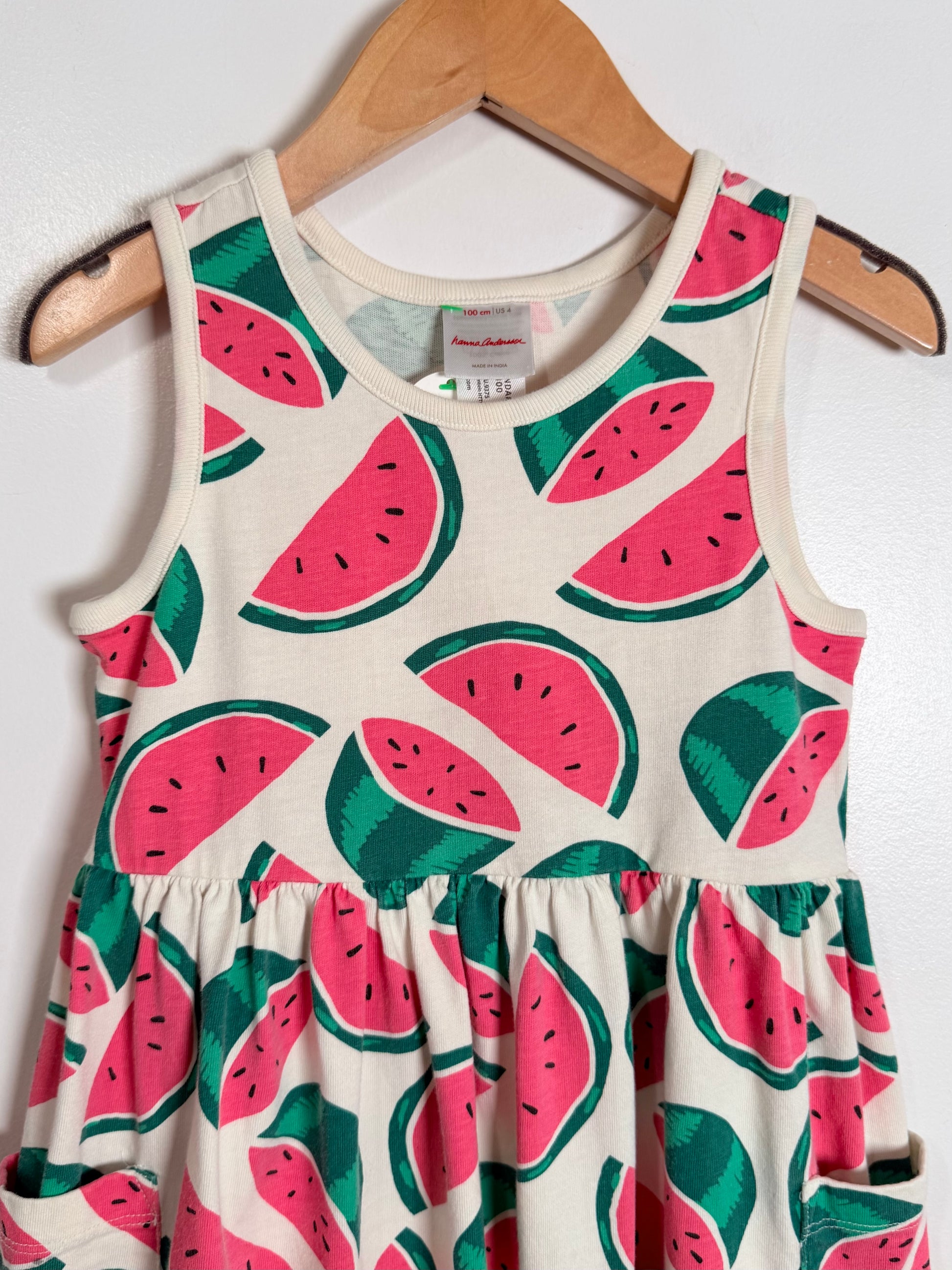 A sleeveless baby dress with a watermelon print, hanging on a wooden hanger against a white wall. The dress features pink watermelon slices with green rinds and black seeds on an off-white background.