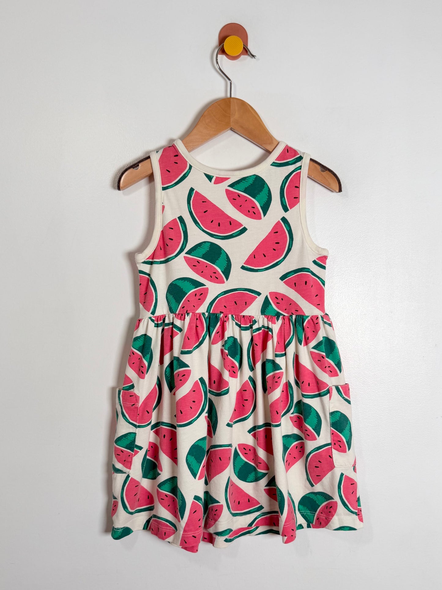 A sleeveless white dress with a watermelon print, hanging on a wooden hanger against a plain white wall.