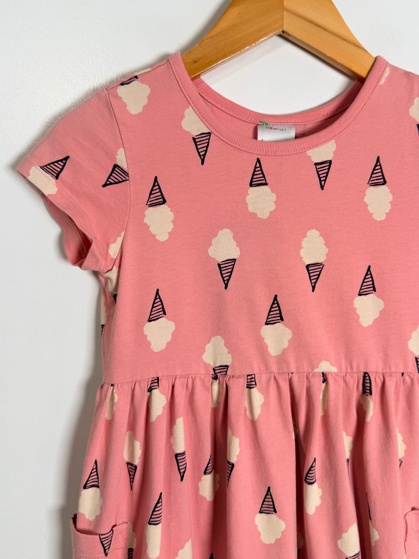 A pink short-sleeve dress with an all-over ice cream cone and cloud print, hanging on a wooden hanger against a plain white wall.