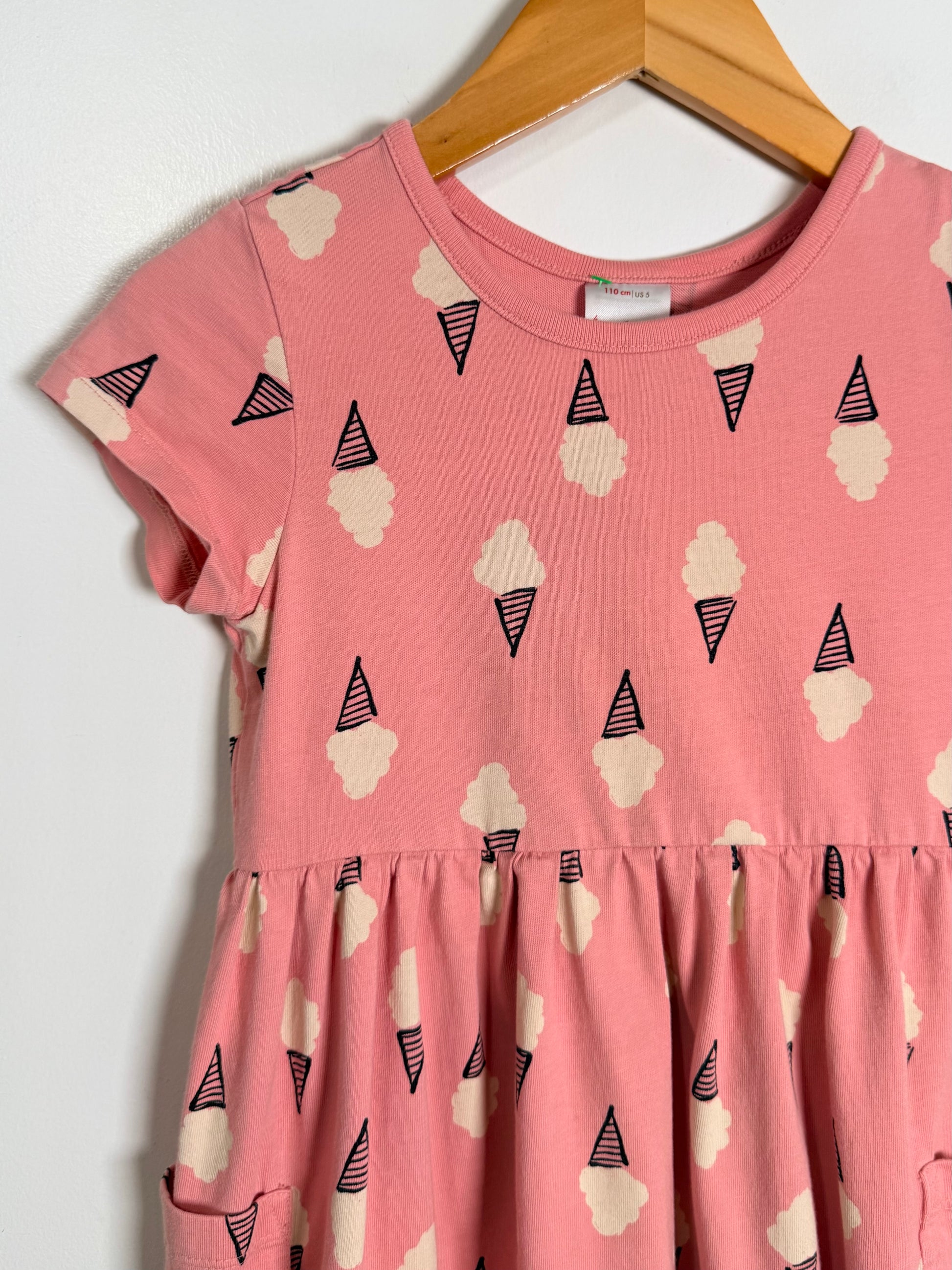A pink short-sleeve dress with an all-over ice cream cone and cloud print, hanging on a wooden hanger against a plain white wall.