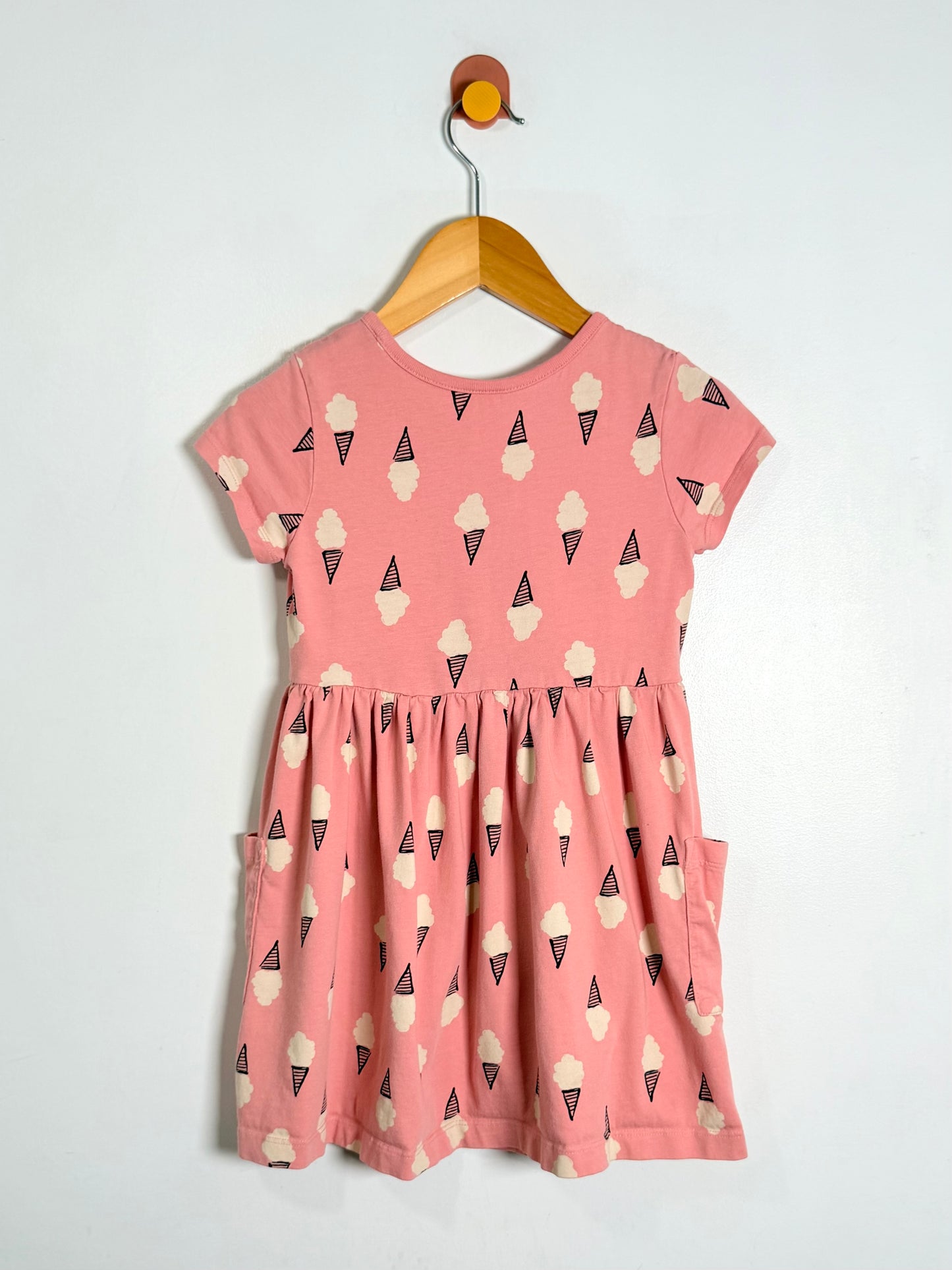 A pink short-sleeve dress with an all-over ice cream cone print, hanging on a wooden hanger against a plain white wall.