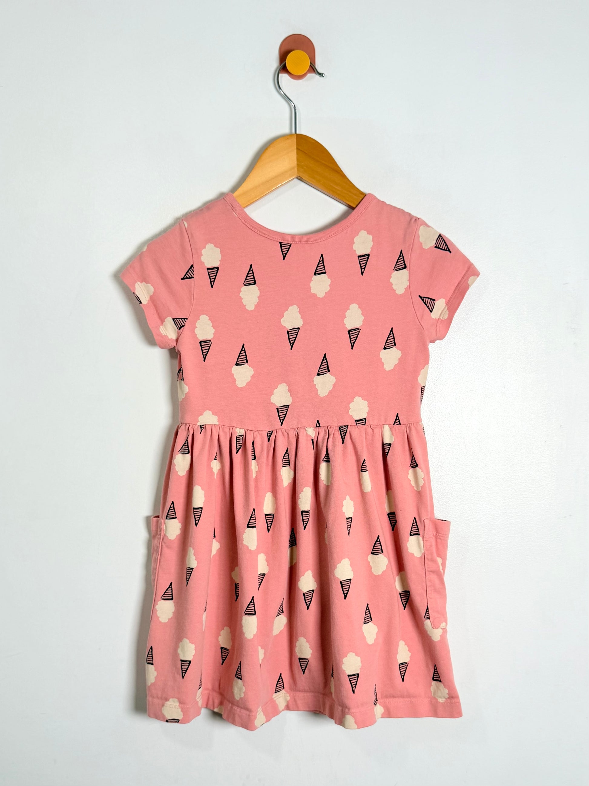 A pink short-sleeve dress with an all-over ice cream cone print, hanging on a wooden hanger against a plain white wall.