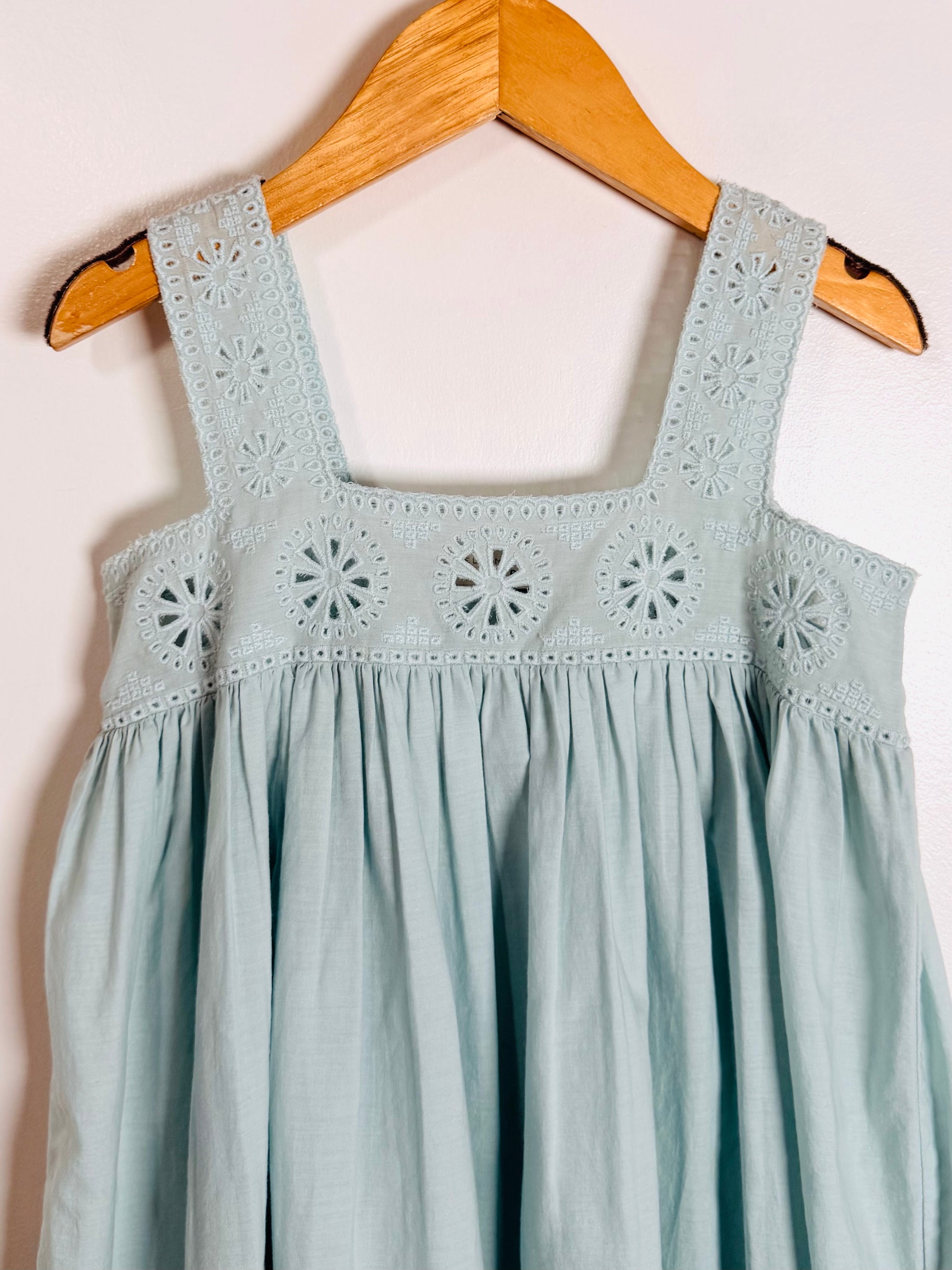 A light blue sleeveless dress with embroidered eyelet detailing on the bodice and straps, hanging on a wooden hanger against a plain white wall.