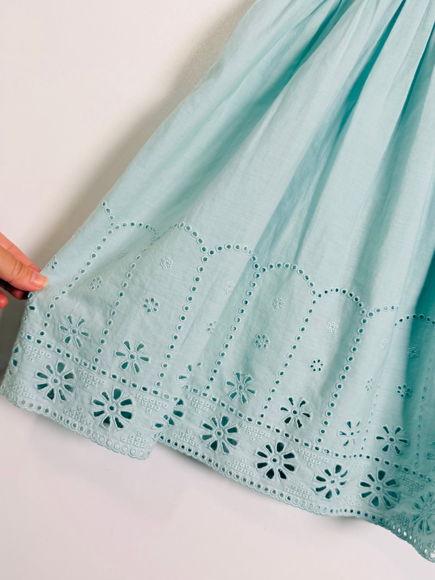 A close-up of a light mint green skirt with intricate eyelet embroidery and scalloped borders, held by a hand on the left side against a plain white background.