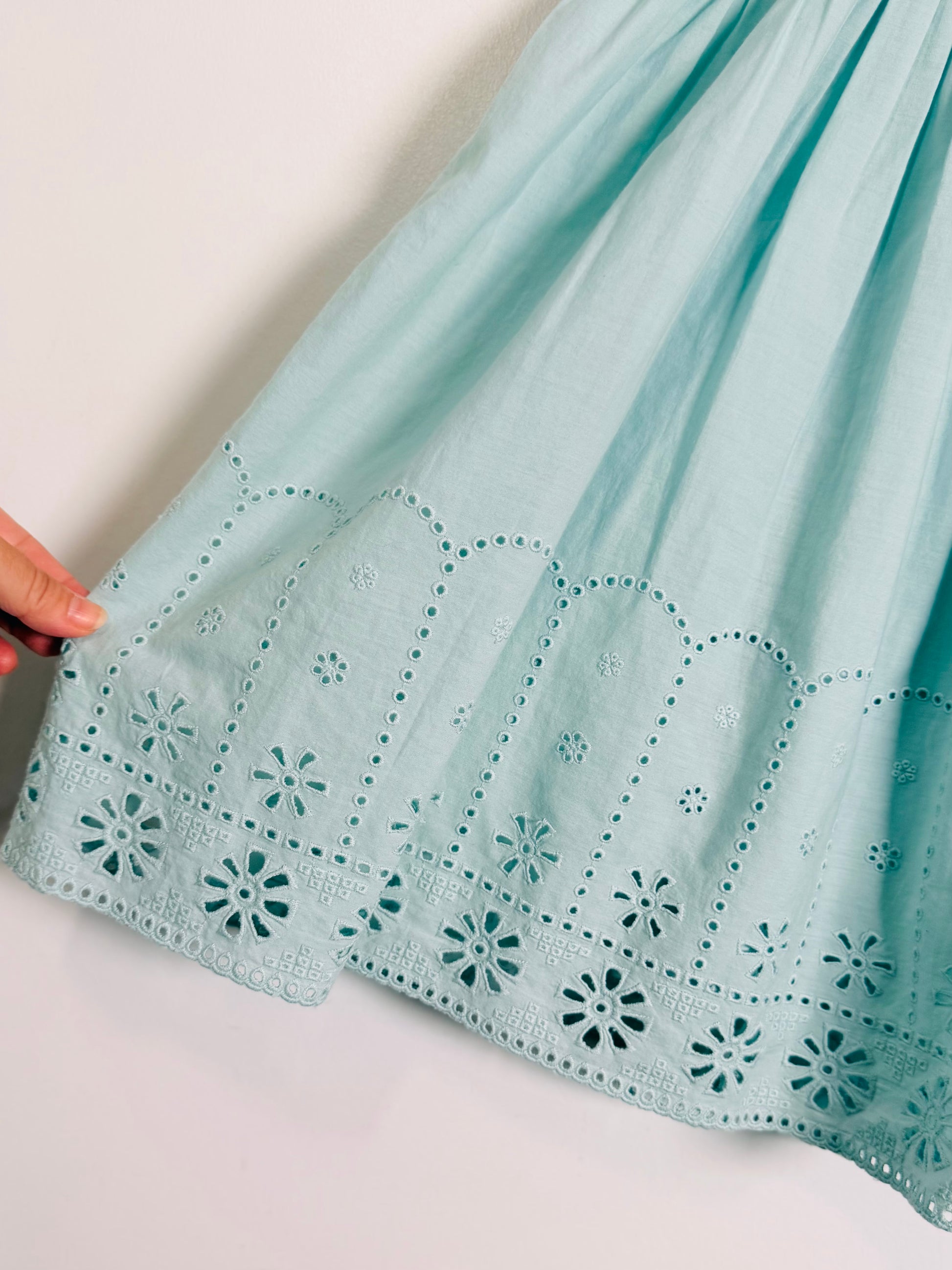 A close-up of a light mint green skirt with intricate eyelet embroidery and scalloped borders, held by a hand on the left side against a plain white background.