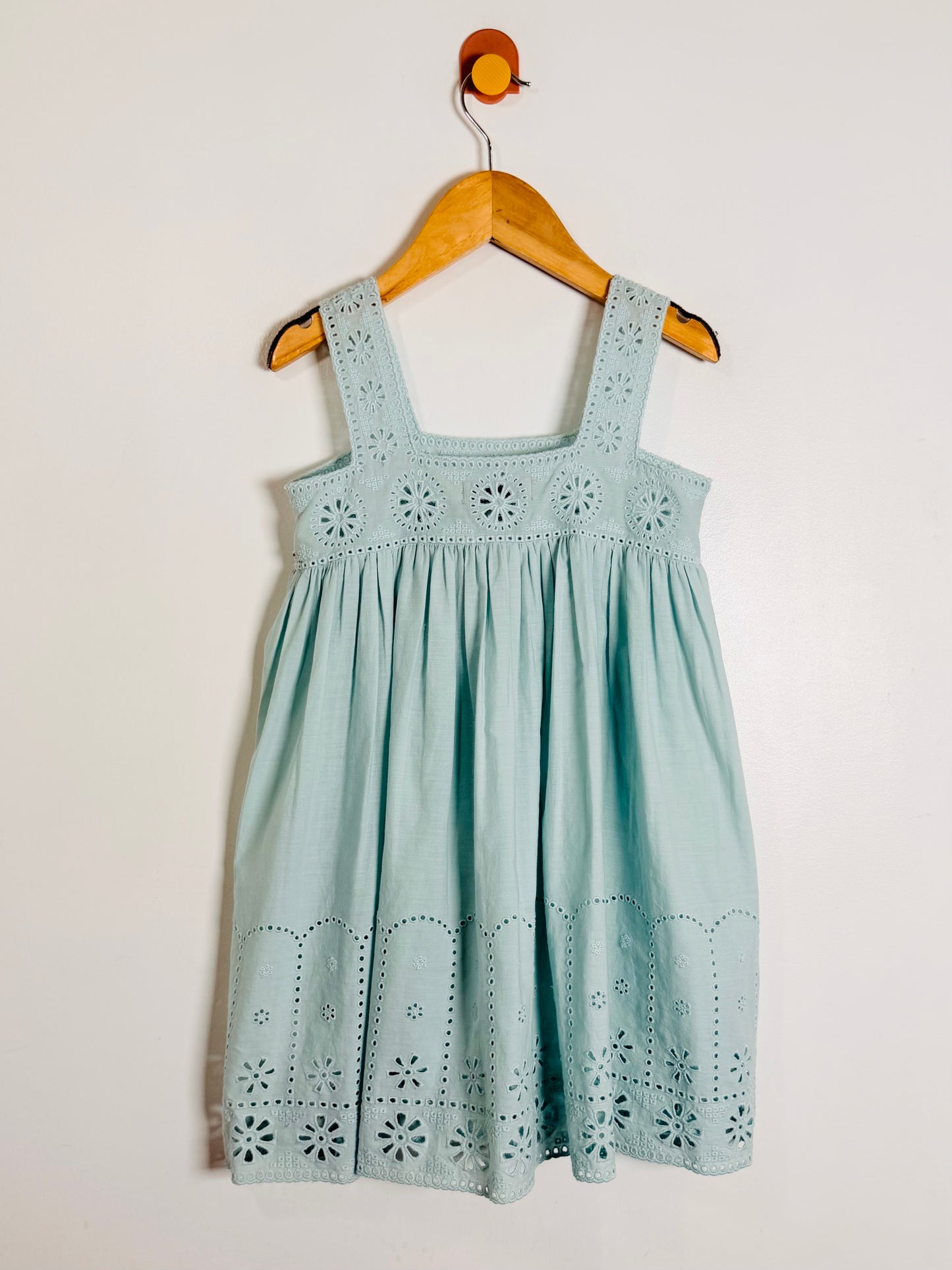 A light blue-green sleeveless dress with eyelet embroidery, hanging on a wooden hanger against a plain off-white wall.
