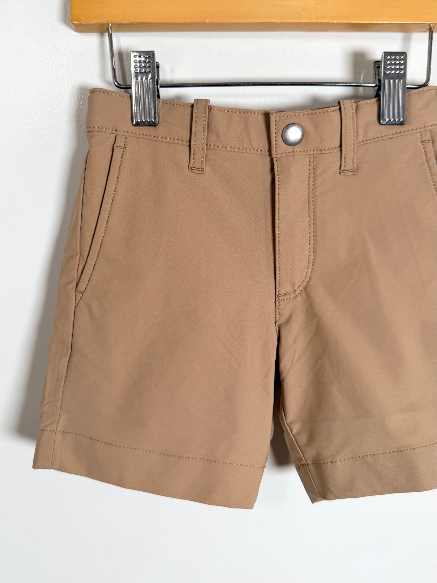 A pair of tan chino shorts hanging from a metal hanger on a wooden hanger bar against a white wall.
