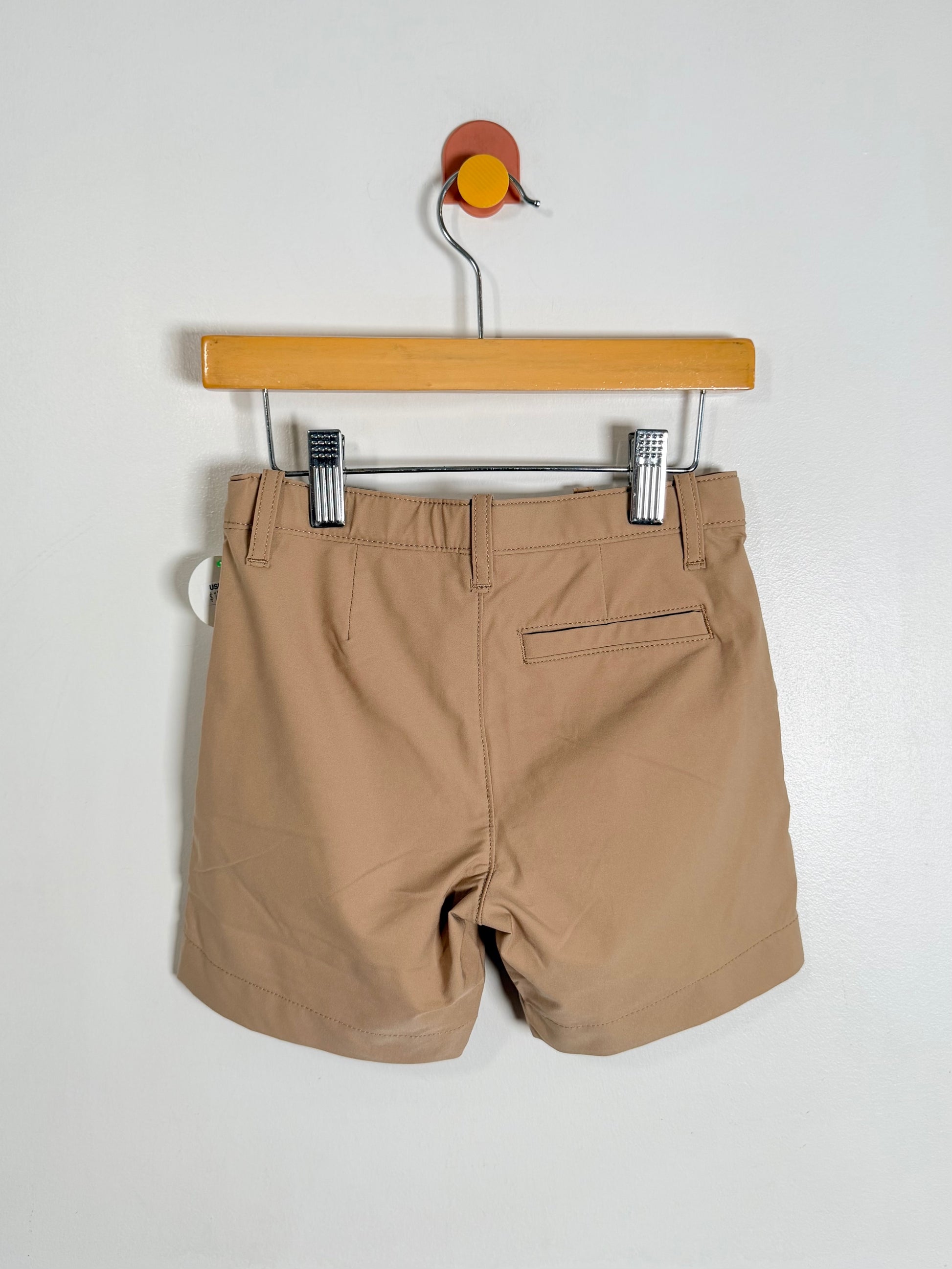 A pair of tan chino shorts hanging from a wooden hanger with two black-and-white striped clips against a plain white wall.