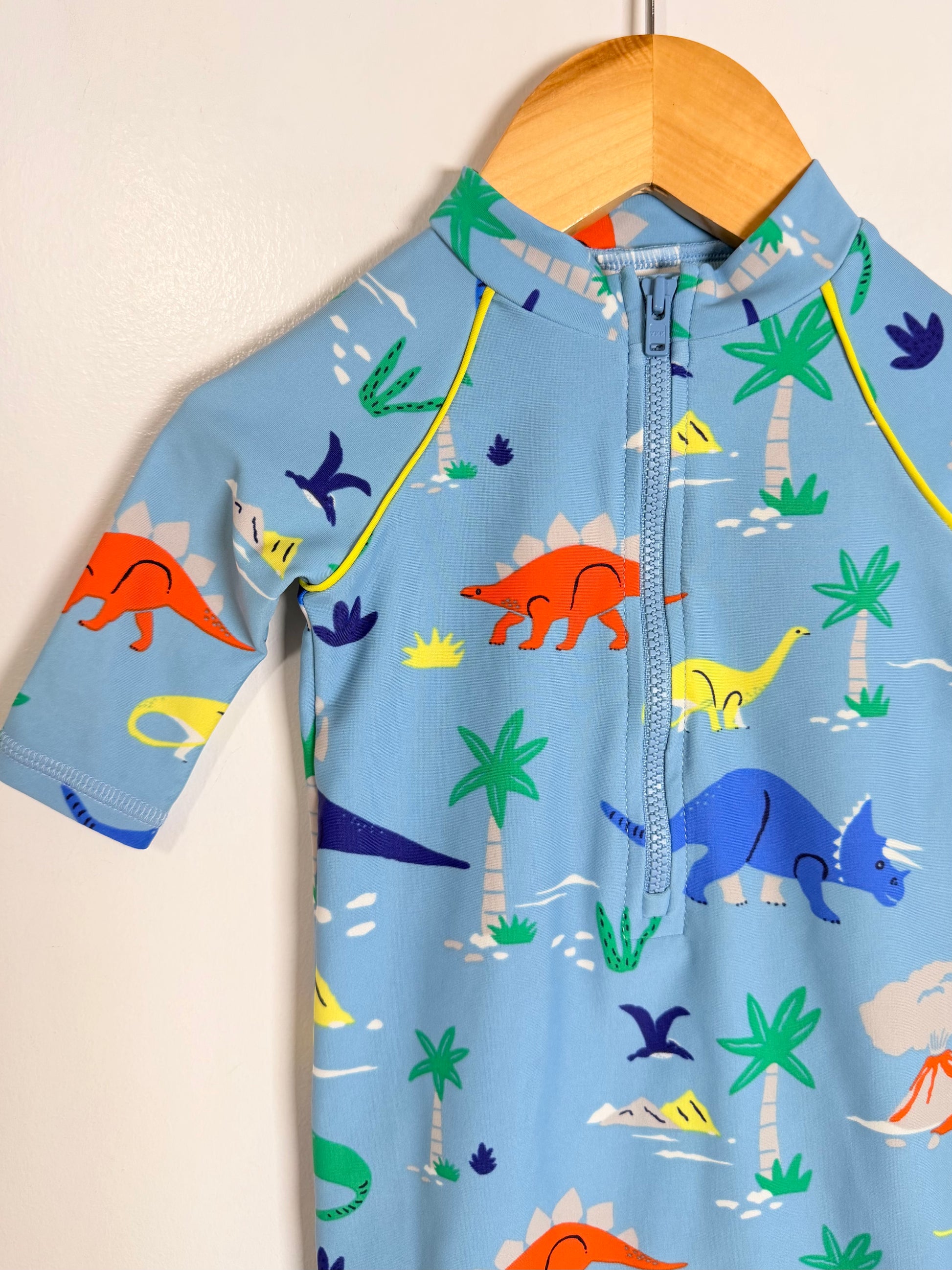 A light blue short-sleeve children's one-piece swimsuit or rash guard with a half-zip neckline, featuring a colorful dinosaur and palm tree print, hanging on a wooden hanger against a plain beige wall.
