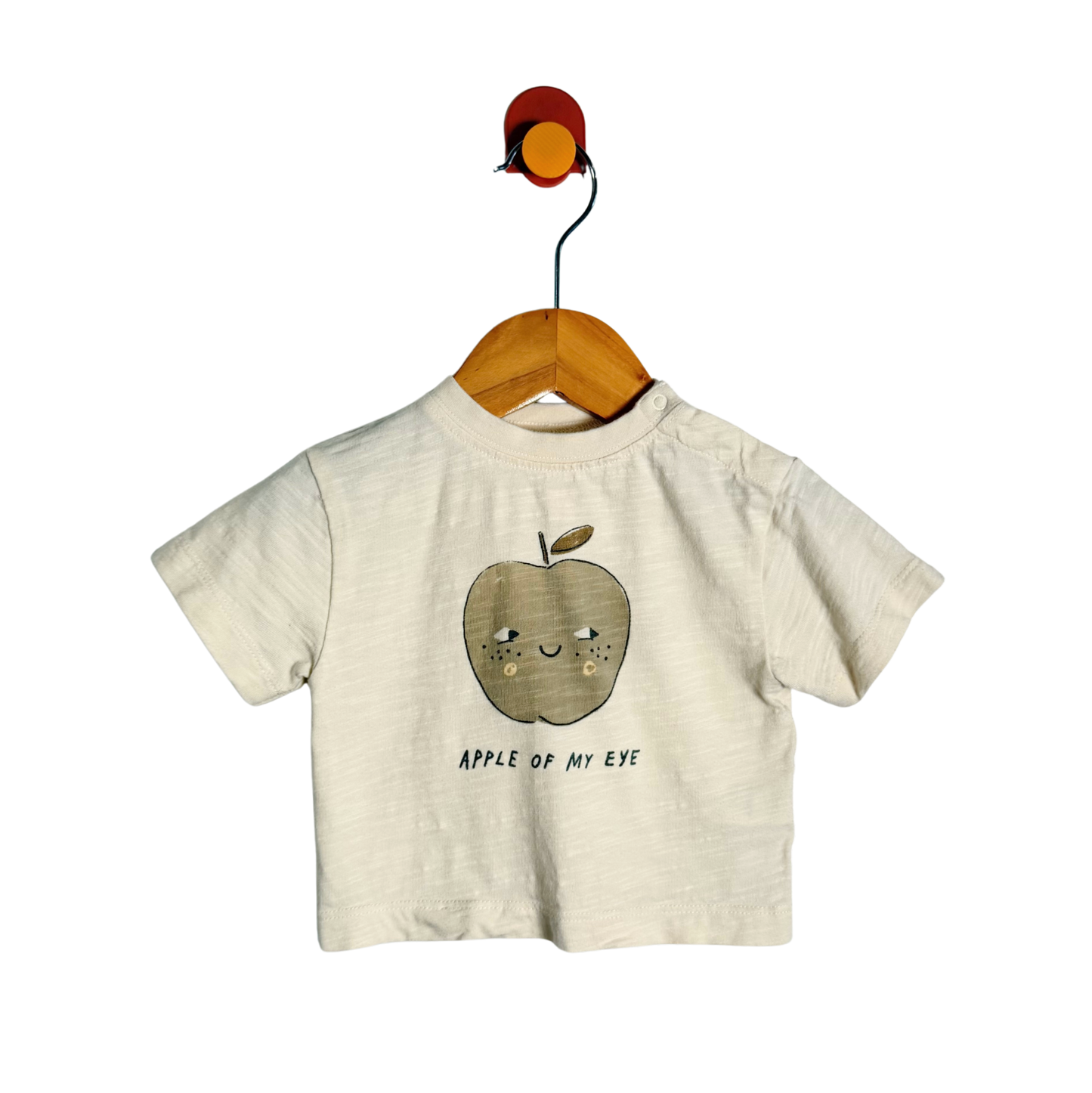 A short-sleeve baby t-shirt in a light cream/off-white textured fabric, featuring a cute cartoon apple graphic with a smiling face and the text 'APPLE OF MY EYE' printed below it. The shirt is displayed on a wooden hanger hanging from a colorful wall hook against a solid white background.