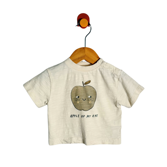 A short-sleeve baby t-shirt in a light cream/off-white textured fabric, featuring a cute cartoon apple graphic with a smiling face and the text 'APPLE OF MY EYE' printed below it. The shirt is displayed on a wooden hanger hanging from a colorful wall hook against a solid white background.