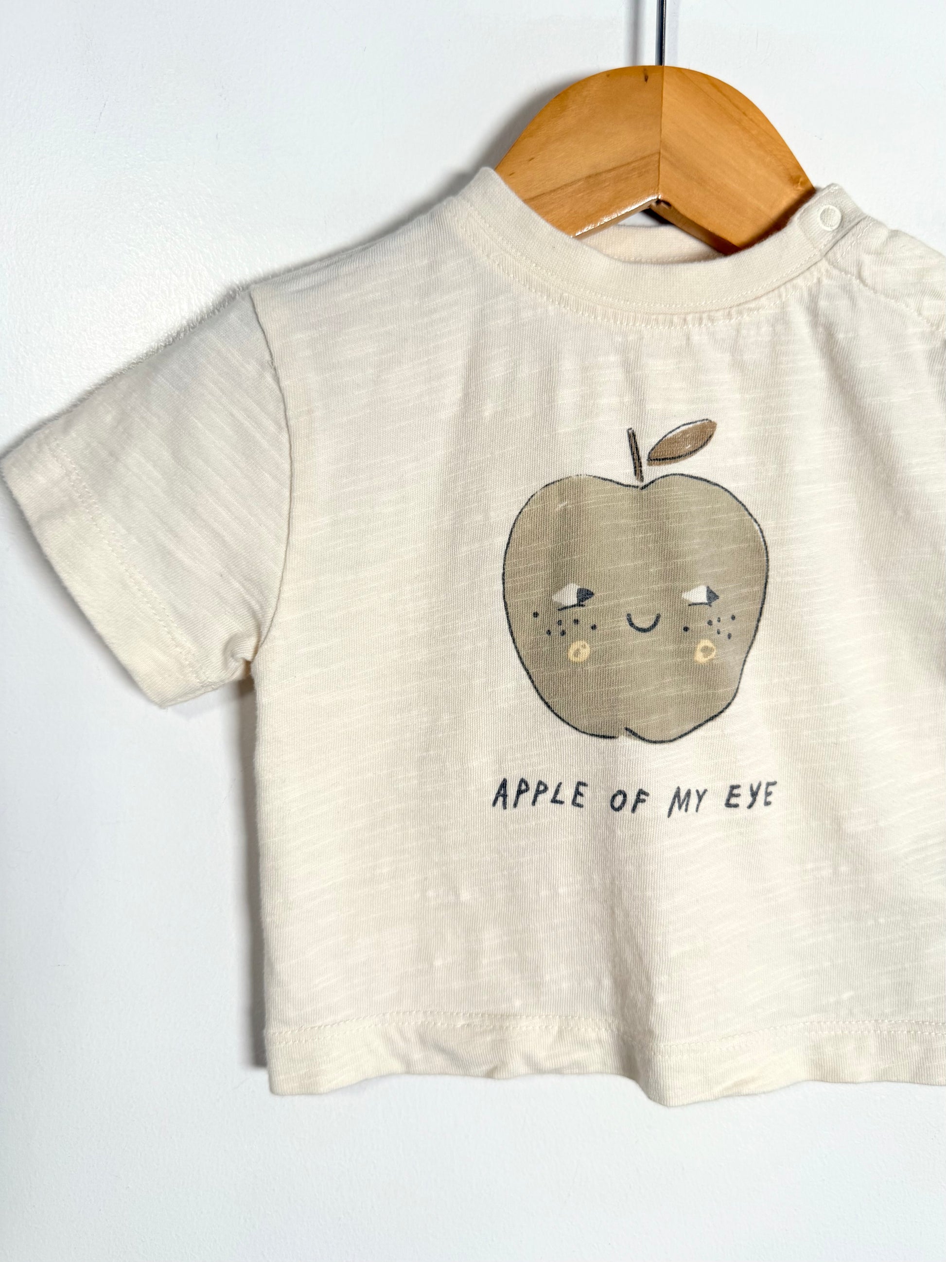 A cream-colored short-sleeve baby t-shirt hanging on a wooden hanger against a white wall. The shirt features a cute embroidered apple graphic with a face and the text 'APPLE OF MY EYE' printed below it.