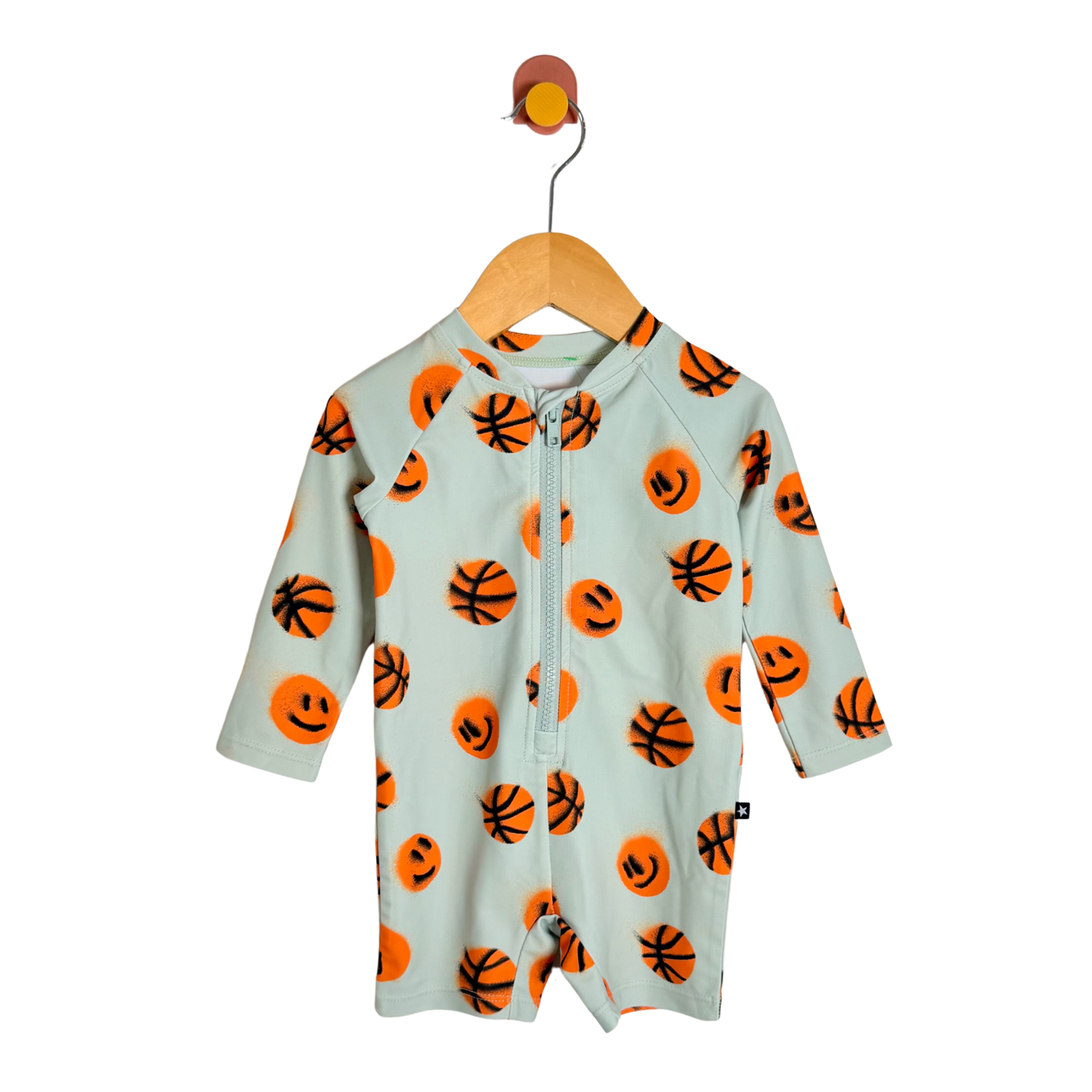 A baby long-sleeve zip-up romper in a pale mint green, decorated with an all-over orange basketball and smiley face pattern, hanging on a wooden hanger against a white background.